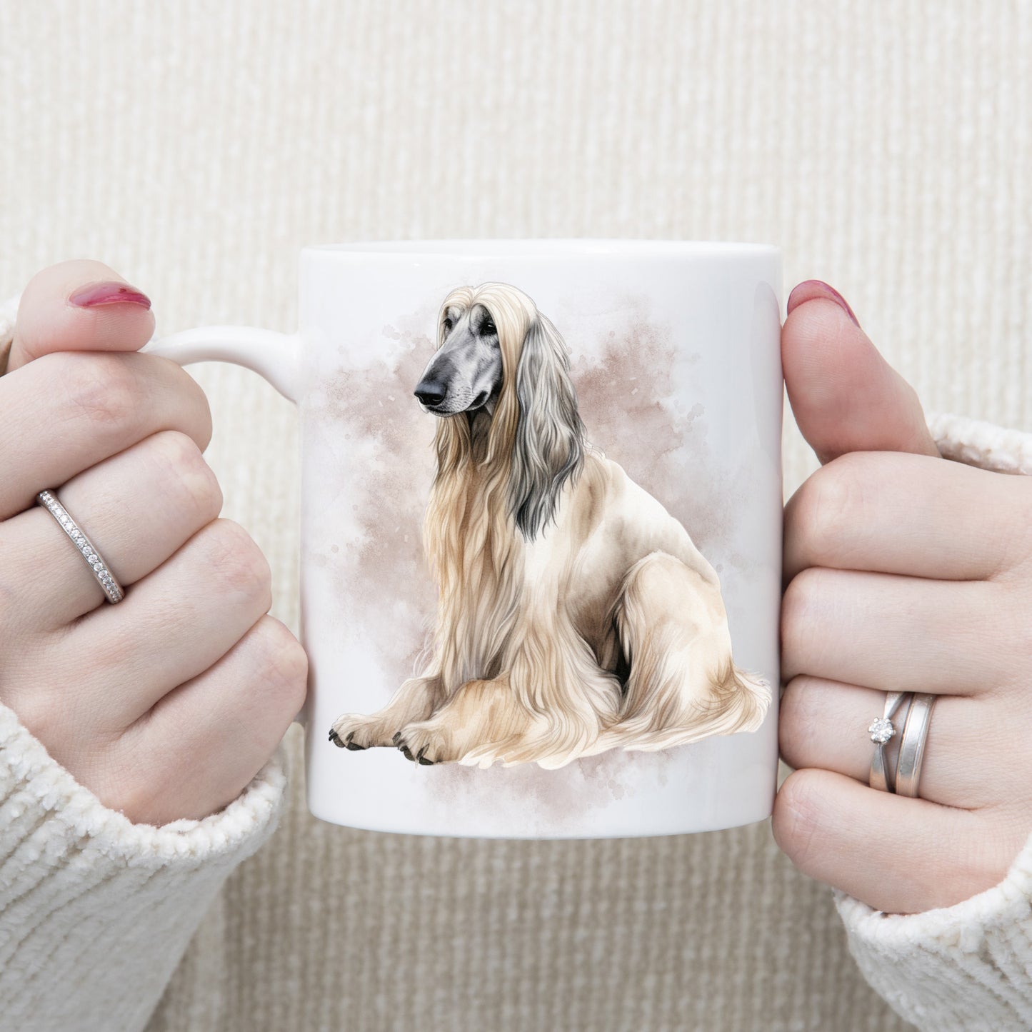 A white ceramic mug with a cream Afghan Hound with long flowing hair sat facing left. A grey/brown smoke effect decorates the background. A woman wearing rings is holding the mug in two hands.