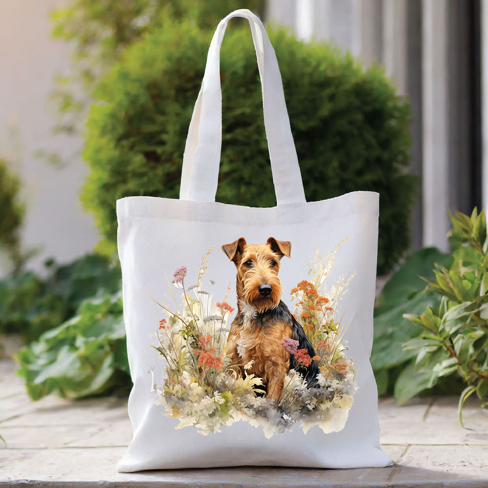 An Airedale Terrier fronts a white tote bag with a long handle. The dog is sat in among orange and purple flowers. The bag is placed in front of a large green shrub with green foliage to the right side.