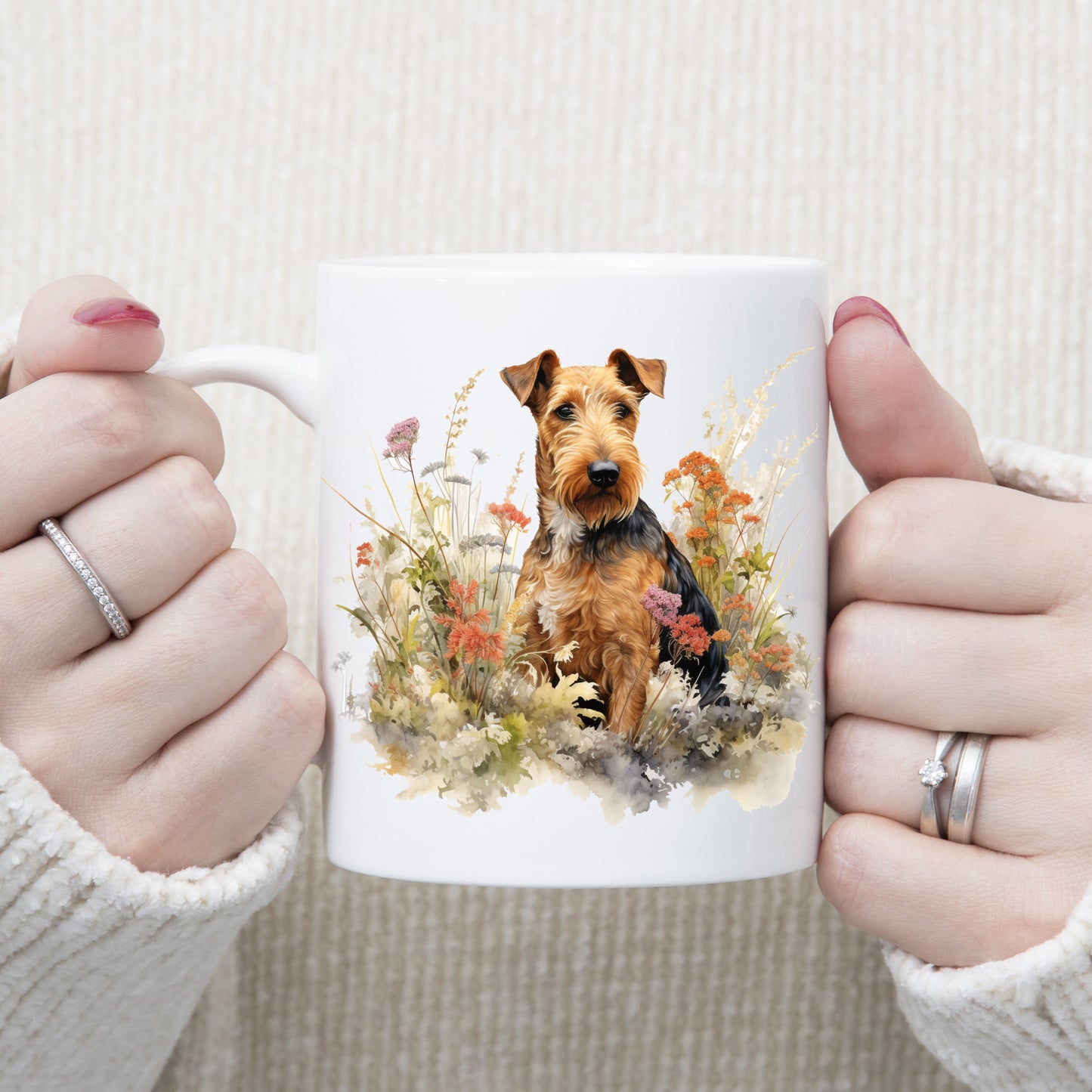 An Airedale Terrier dog is in a bed of orange and purple wildflowers on a white ceramic mug. The mug is being held by a woman with both hands.
