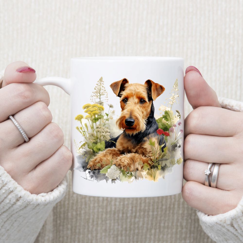 An Airedale Terrier dog is laid in a bed of flowers on a white ceramic mug. A woman is holding the mug in two hands.