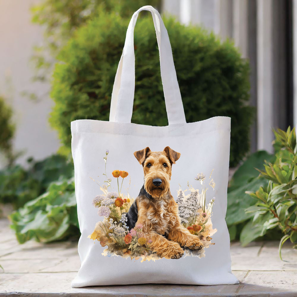 White tote bag with a long handle shows an Airedale Terrier laid among a flower bed and looking forward. The bag is placed in front of a large green shrub with green foliage to the right side.