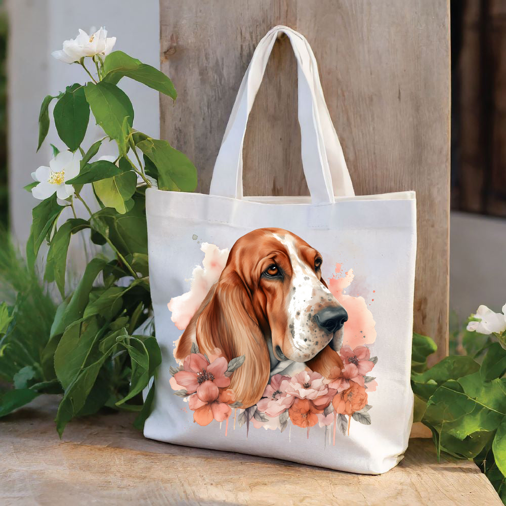 White tote bag with a long handle shows a red and white Basset Hound head shot placed among pink and peach flowers and looking right. The bag is stood against a wood panel. Green foliage and white flowers decorate.