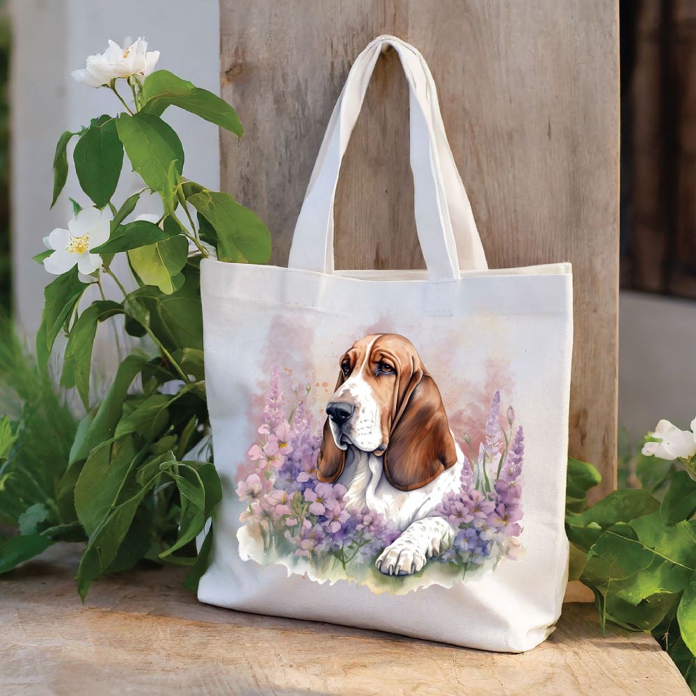 White tote bag with a long handle shows a red and white Basset Hound laid among a purple flower bed and looking toward the left. The bag is stood against a wood panel. Green foliage and white flowers decorate .