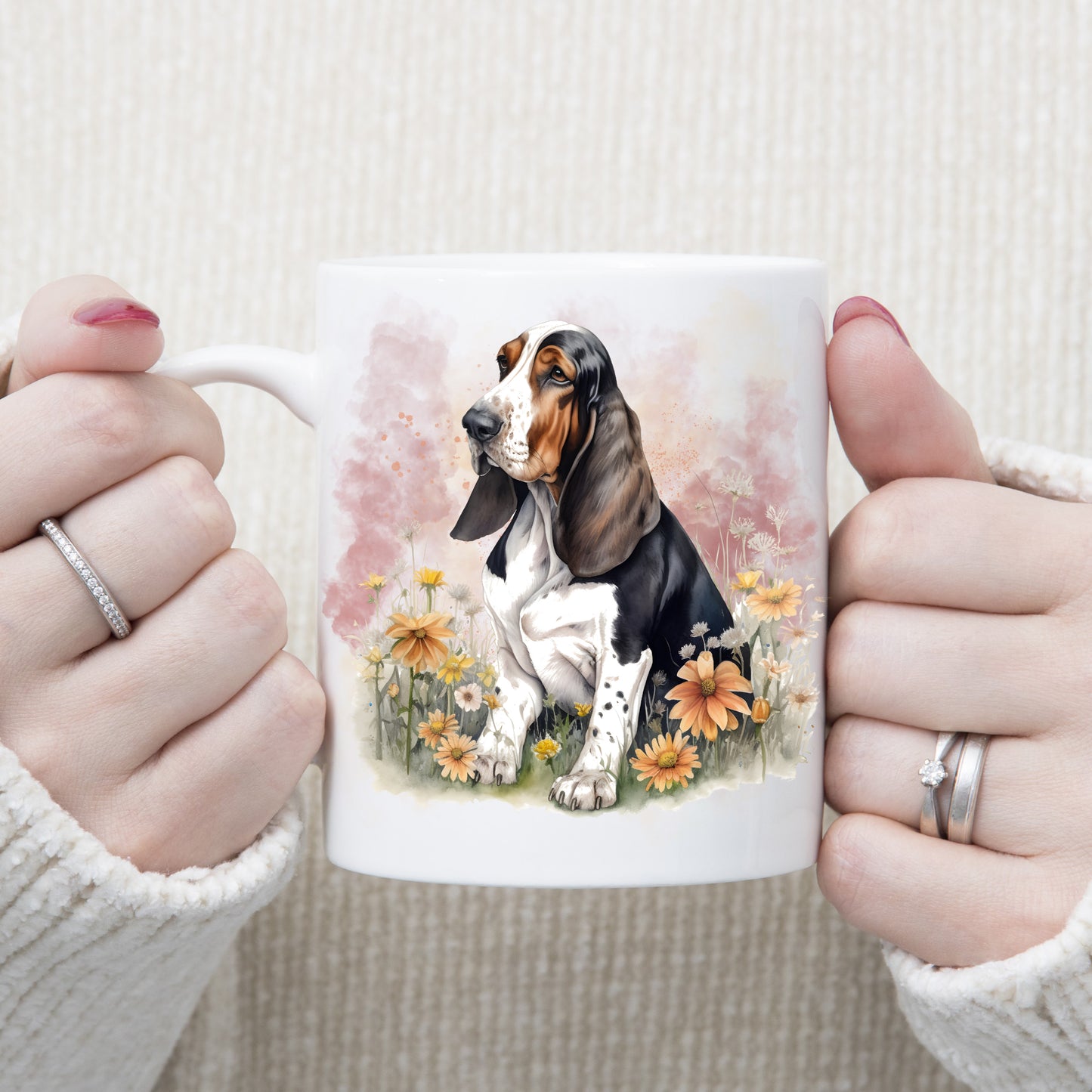 A tri-colour Basset Hound sits facing left among orange flowers. A pink smoky background decorates this white ceramic mug. The mug which is being held by a woman with both hands.