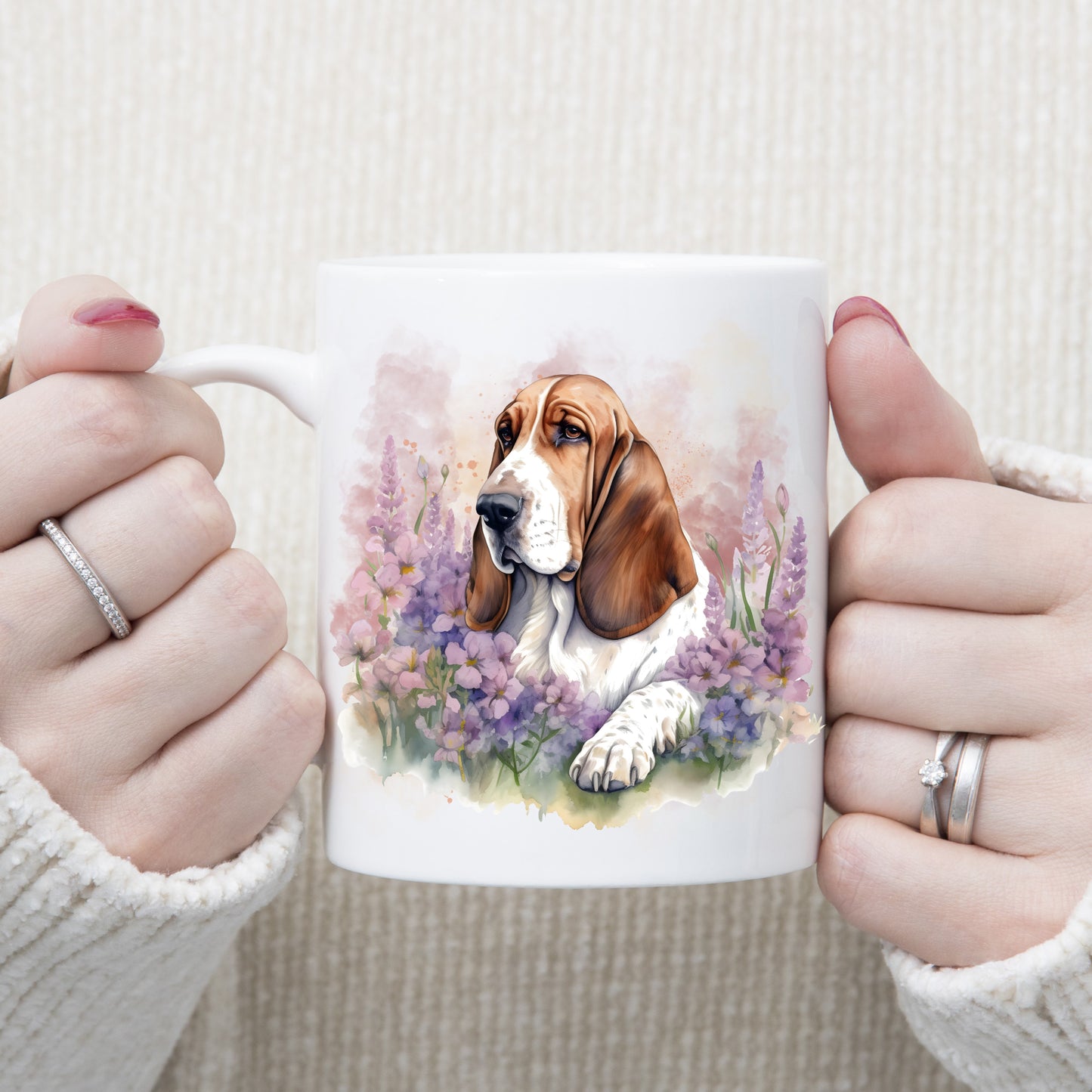 A red and white Basset Hound is laid among purple flowers on a white ceramic mug. A pink smoke effect decorates the background. A woman is holding the mug in two hands.