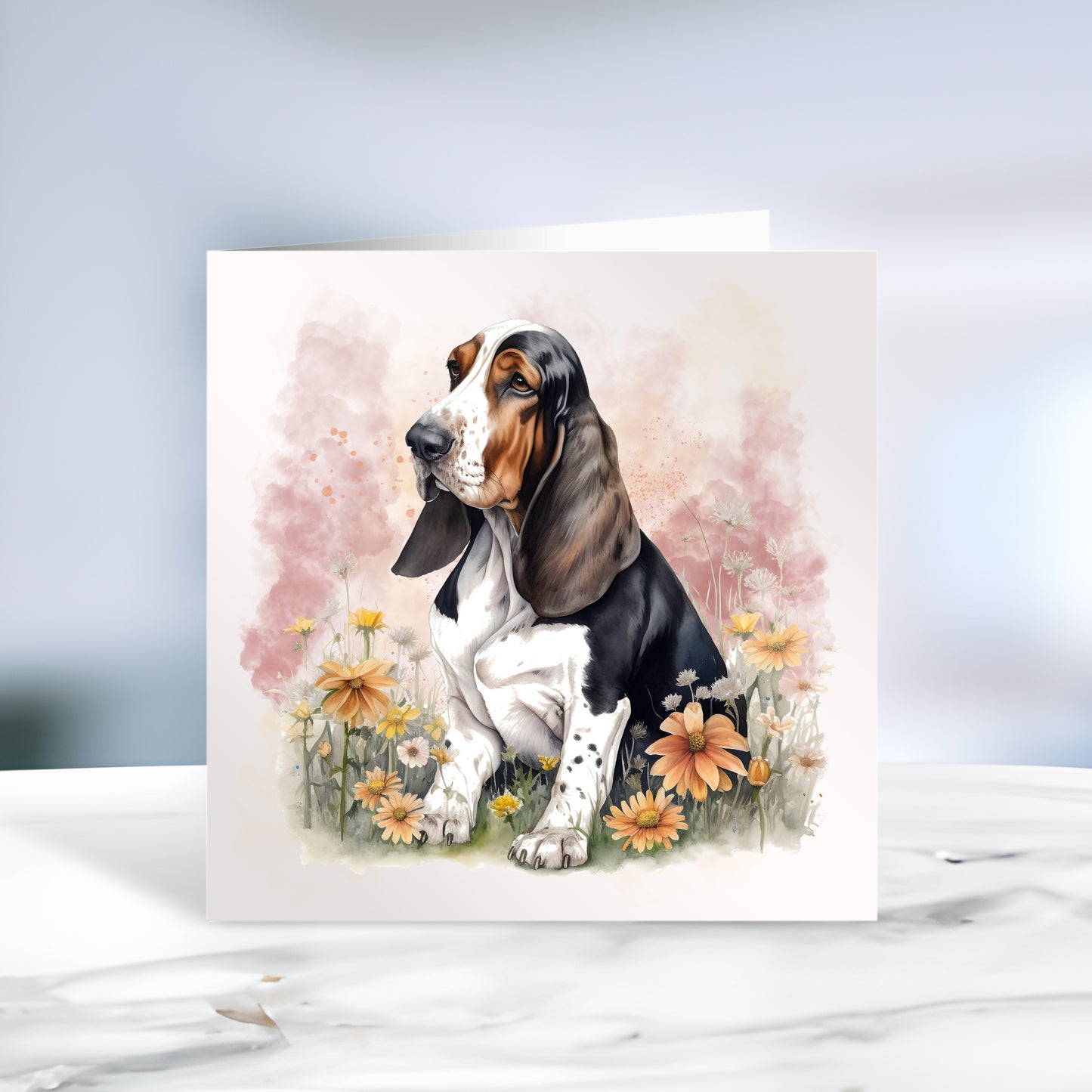 Basset Hound sat among flowers and facing left on a white card or notelet with a smokey pink background. Card is sitting on a marble surface.