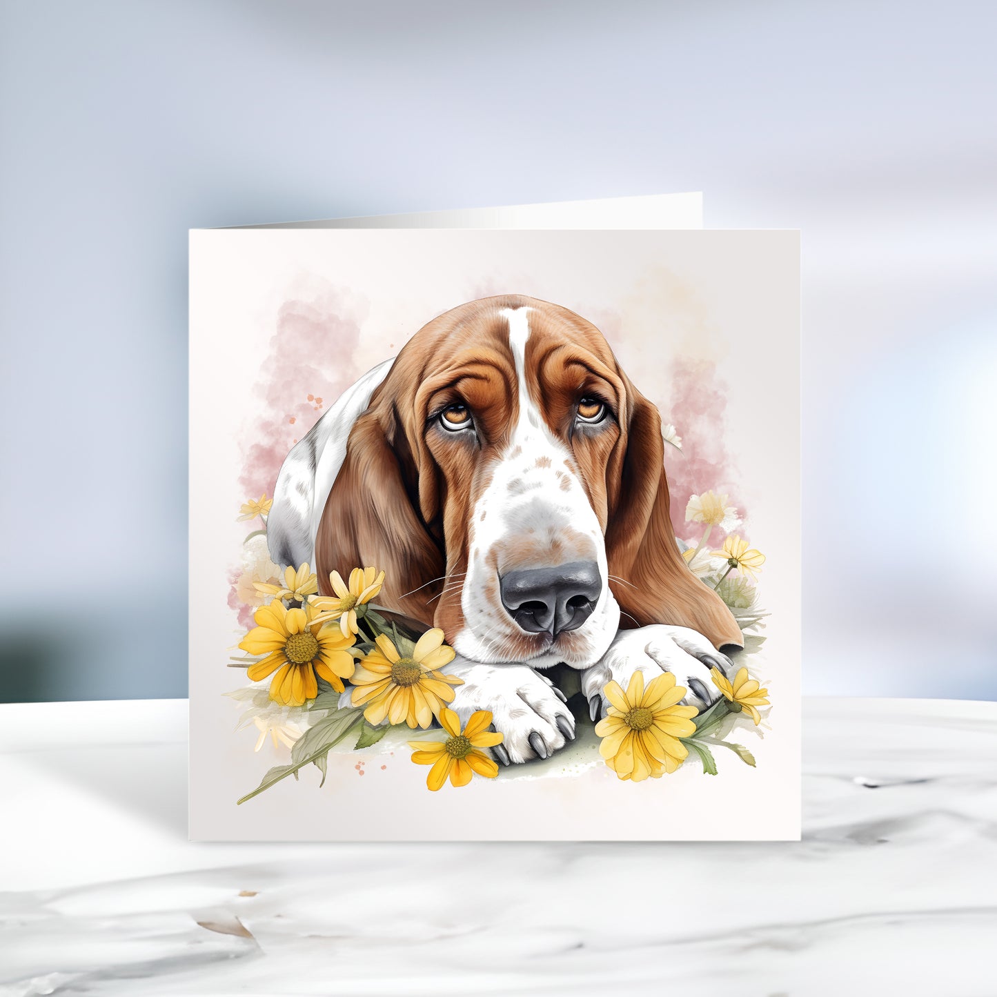 Basset Hound laid among yellow flowers and facing forward on a white card or notelet with a smokey pink background. Card is sitting on a marble surface.