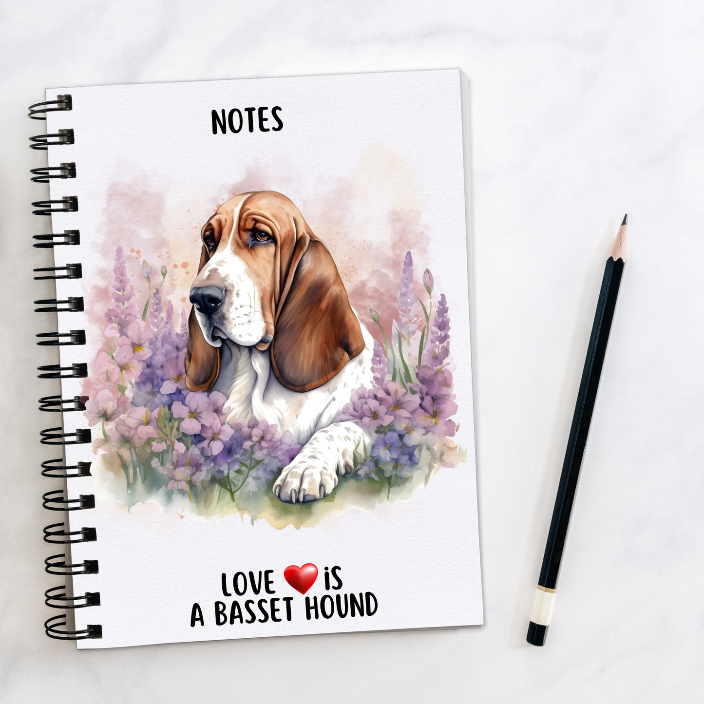 Basset Hound laid among purple flowers and facing left on a white notebook. A pencil is to the right hand side.