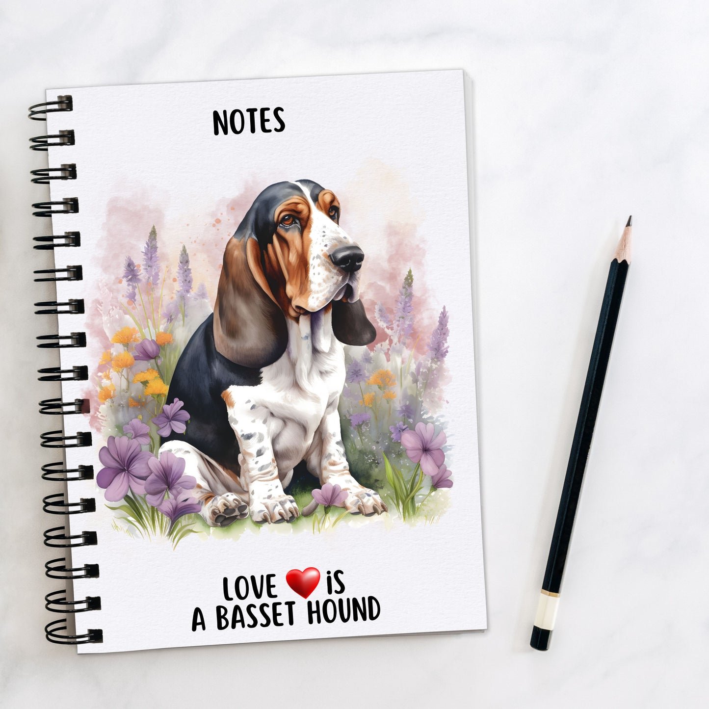Basset Hound sat among purple flowers and facing right on a white notebook. A pencil is to the right hand side.
