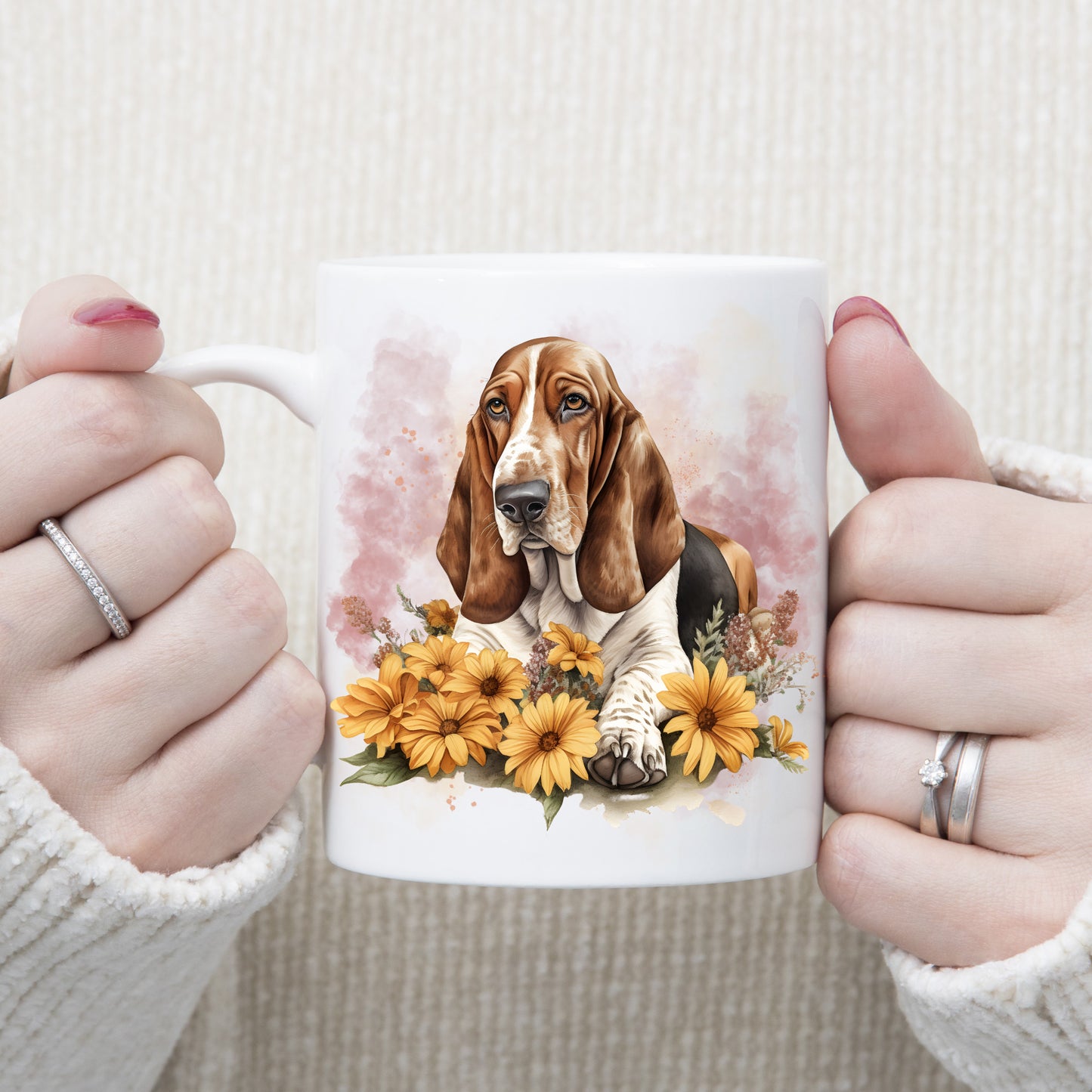 A Basset Hound is laid among orange daisy type flowers on a white ceramic mug. A pink smoke effect decorates the background. A woman is holding the mug in two hands.