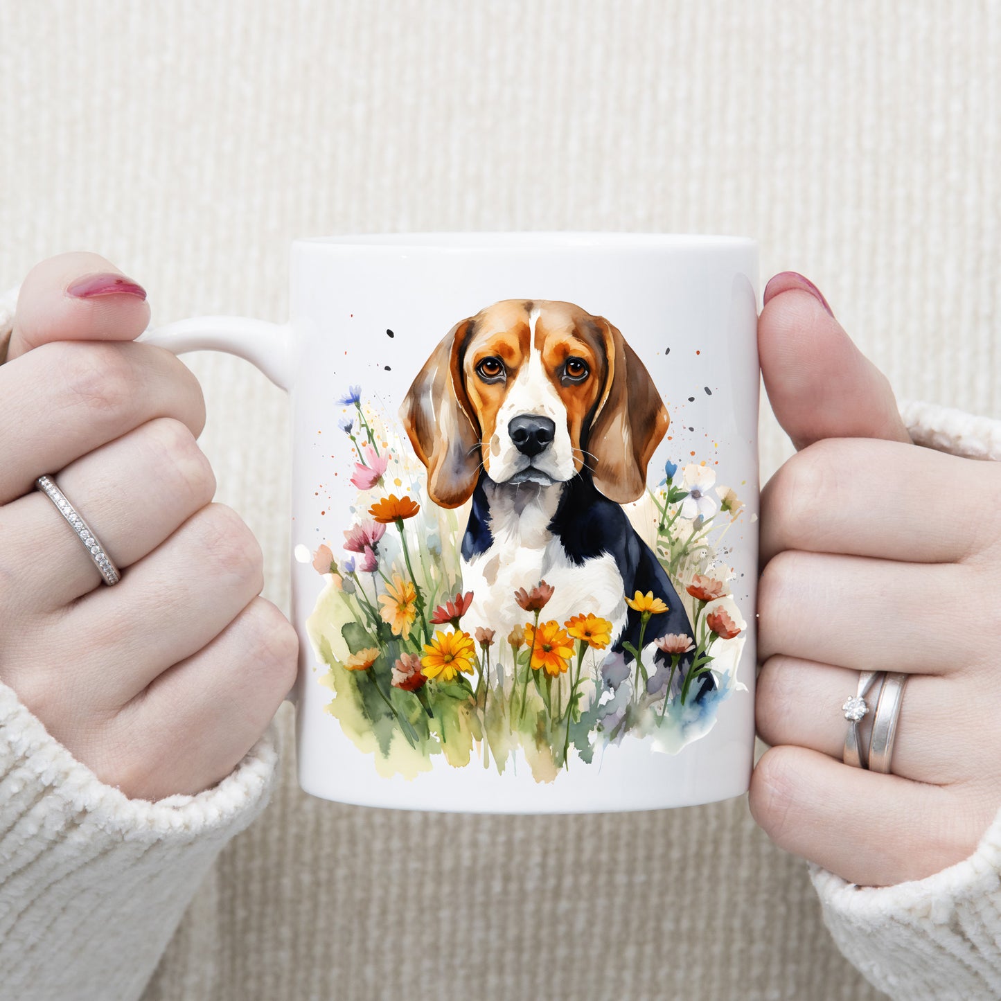 A Beagle dog is sat facing forward among orange daisy-like wildflowers on a white ceramic mug. A woman is holding the mug with both hands.