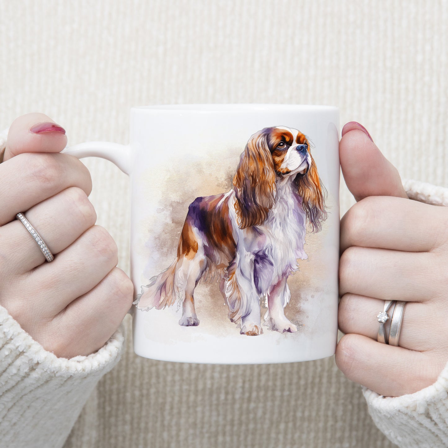 A Blenheim Cavalier King Charles Spaniel is standing in front of a brown and gold misty background on this white ceramic mug. The mug is being held by a woman with both hands.