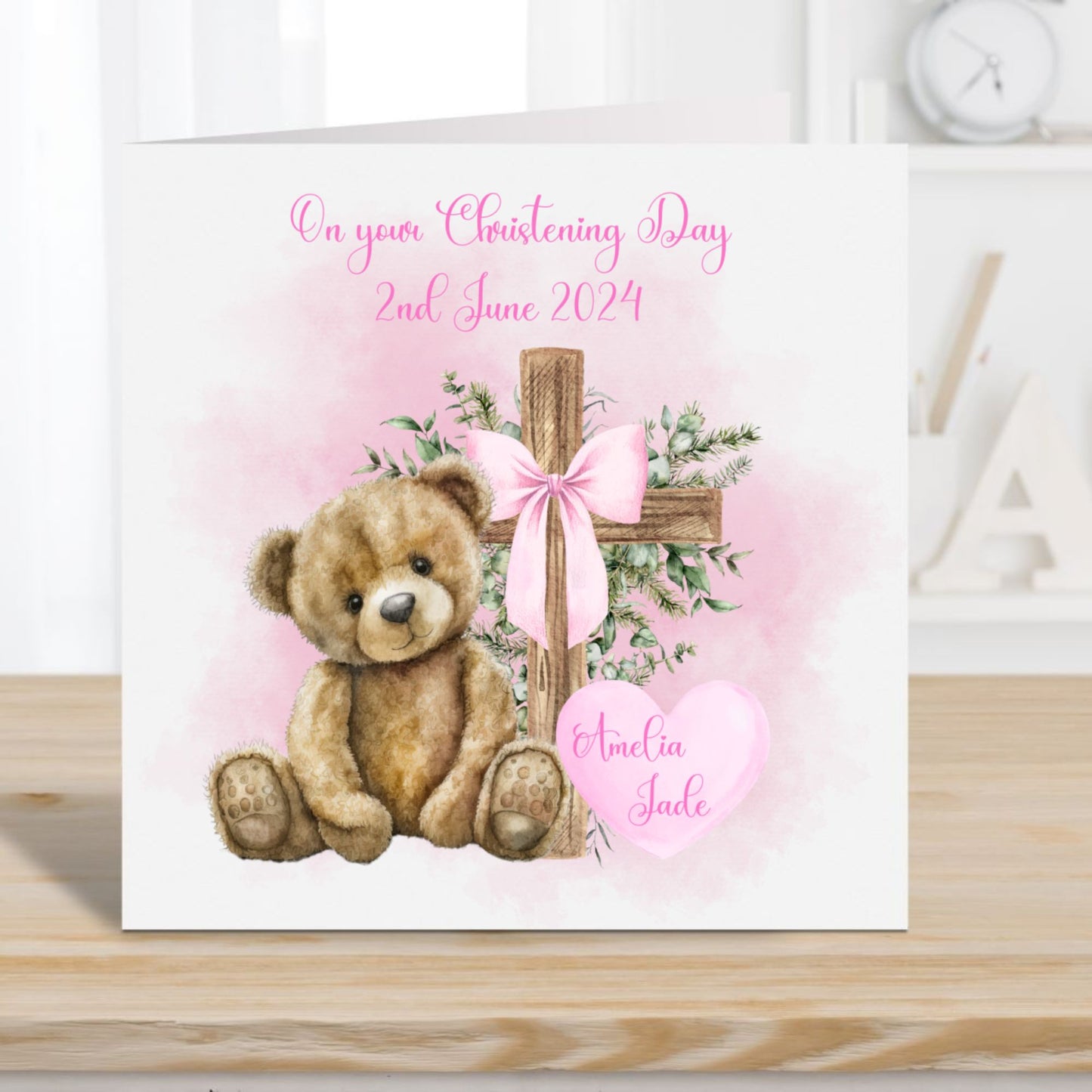 Personalised Christening card for a girl. Teddy bear and cross with a pink ribbon bow. Card placed on a wooden surface with Alphabet letters and a clock in the background.