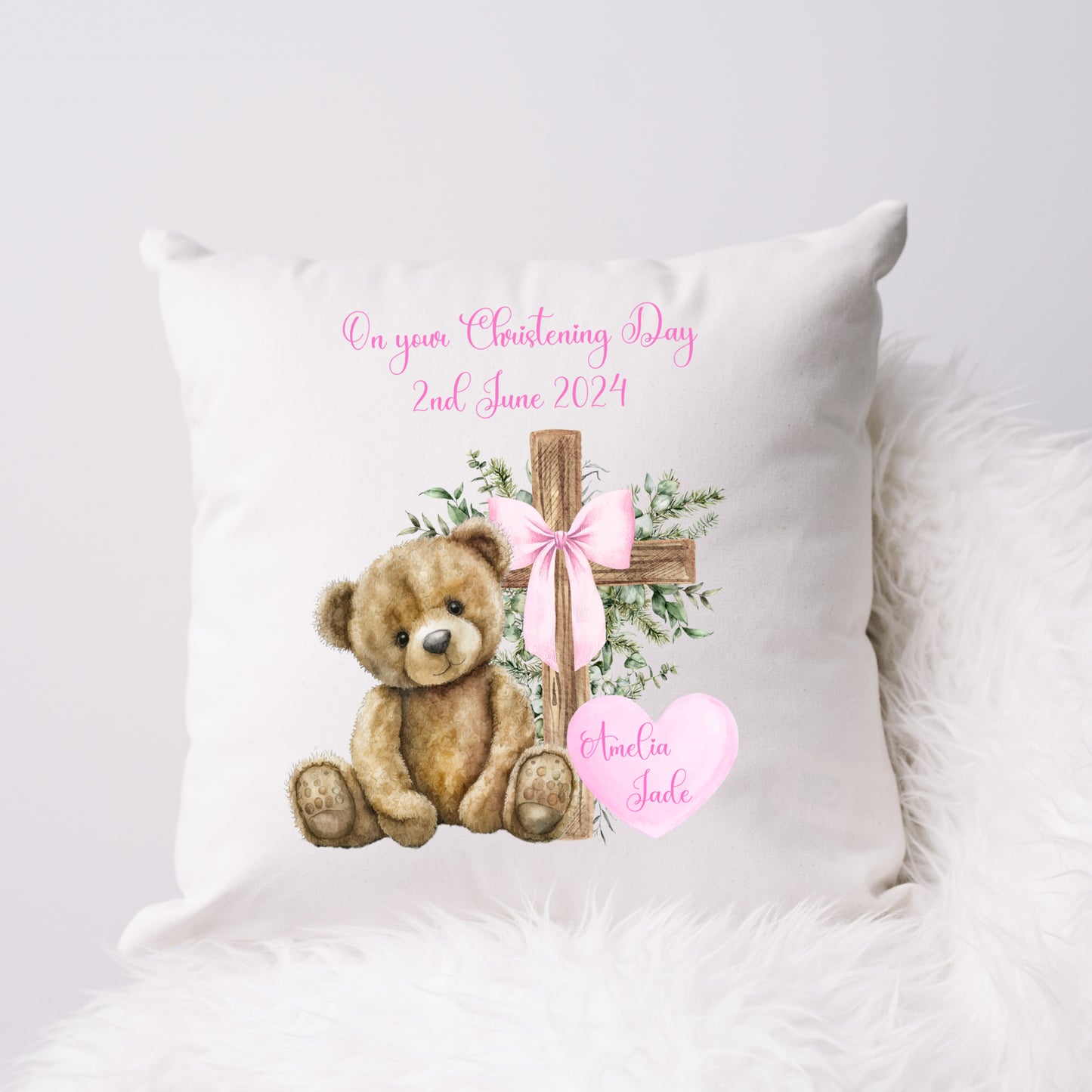 Personalised White Christening Cushion for a girl featuring a teddy bear sitting beside a cross with a pink ribbon bow and green foliage.