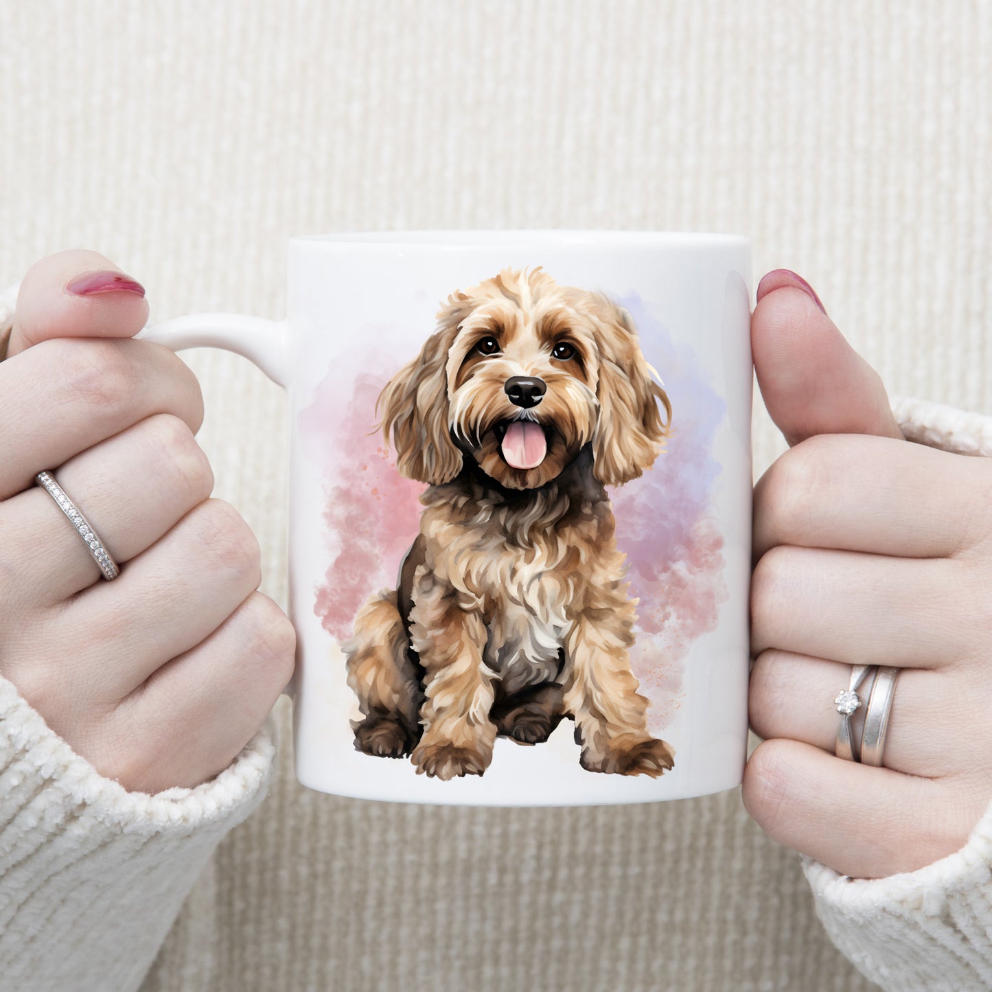 A watercolour image of a cute Cockapoo sitting and looking forward in front of a pink and lilac smoky background, adorns this white ceramic mug.The mug is being held in two hands.