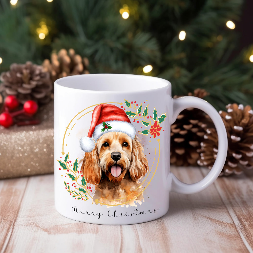A ceramic mug with a watercolour design featuring a Cockapoo dog wearing a Santa hat, positioned in front of a Christmas tree and gifts.