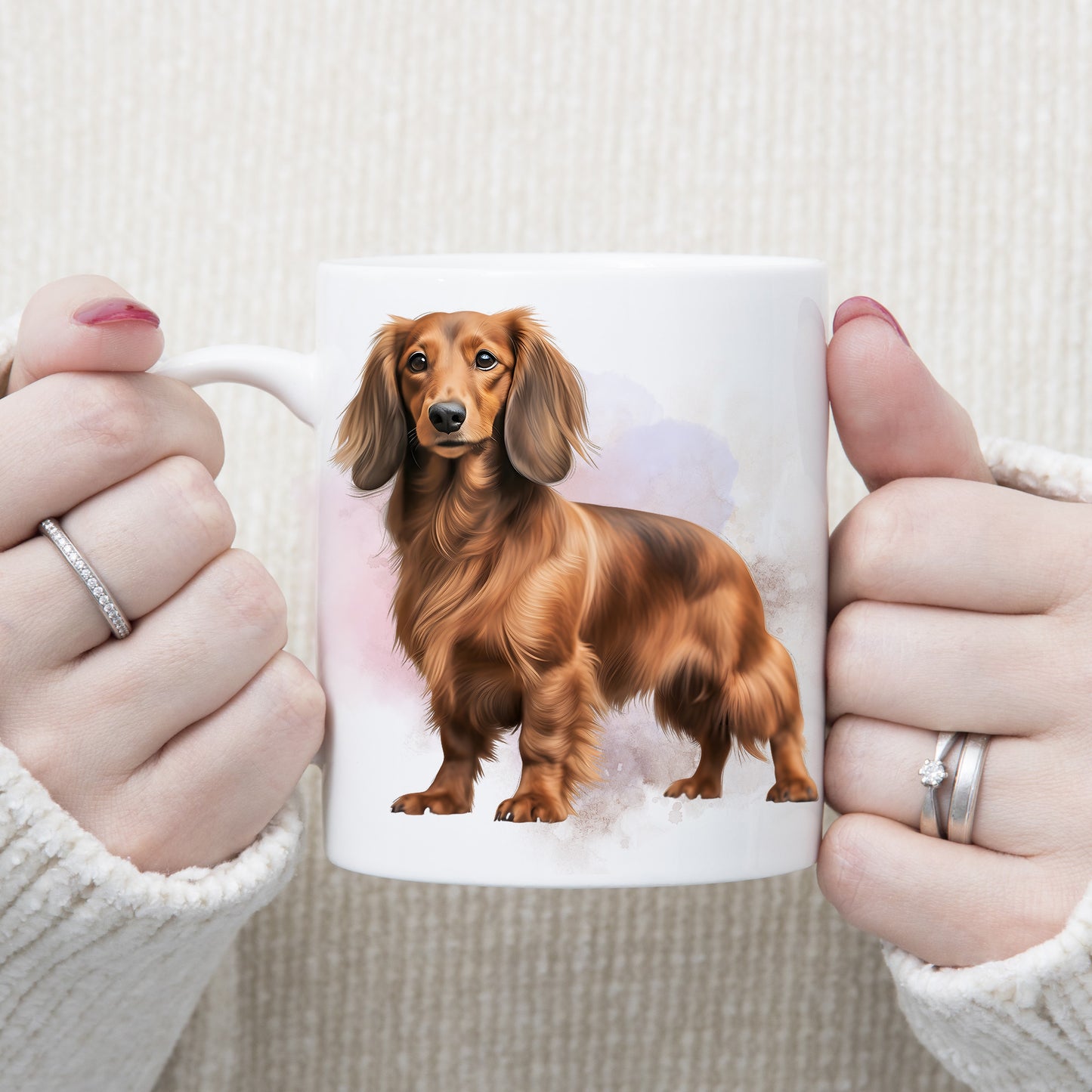 A long haired Dachshund is standing against a lilac and pink background on this white ceramic mug. The mug is being held by a woman with both hands.