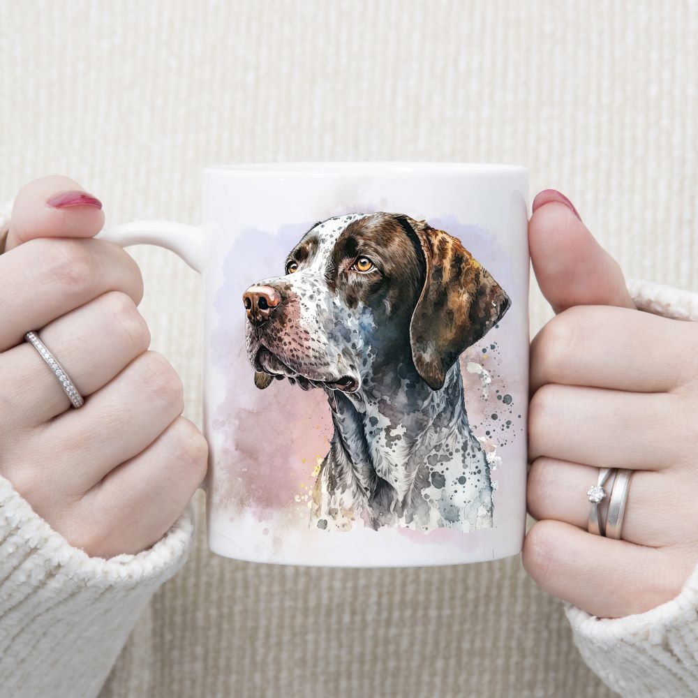 A water-colour image of an English Pointer with a speckly coat on a white ceramic mug. A Lilac and pink smoky effect background with a few splashes decorate. A woman wearing rings is holding the mug in two hands.