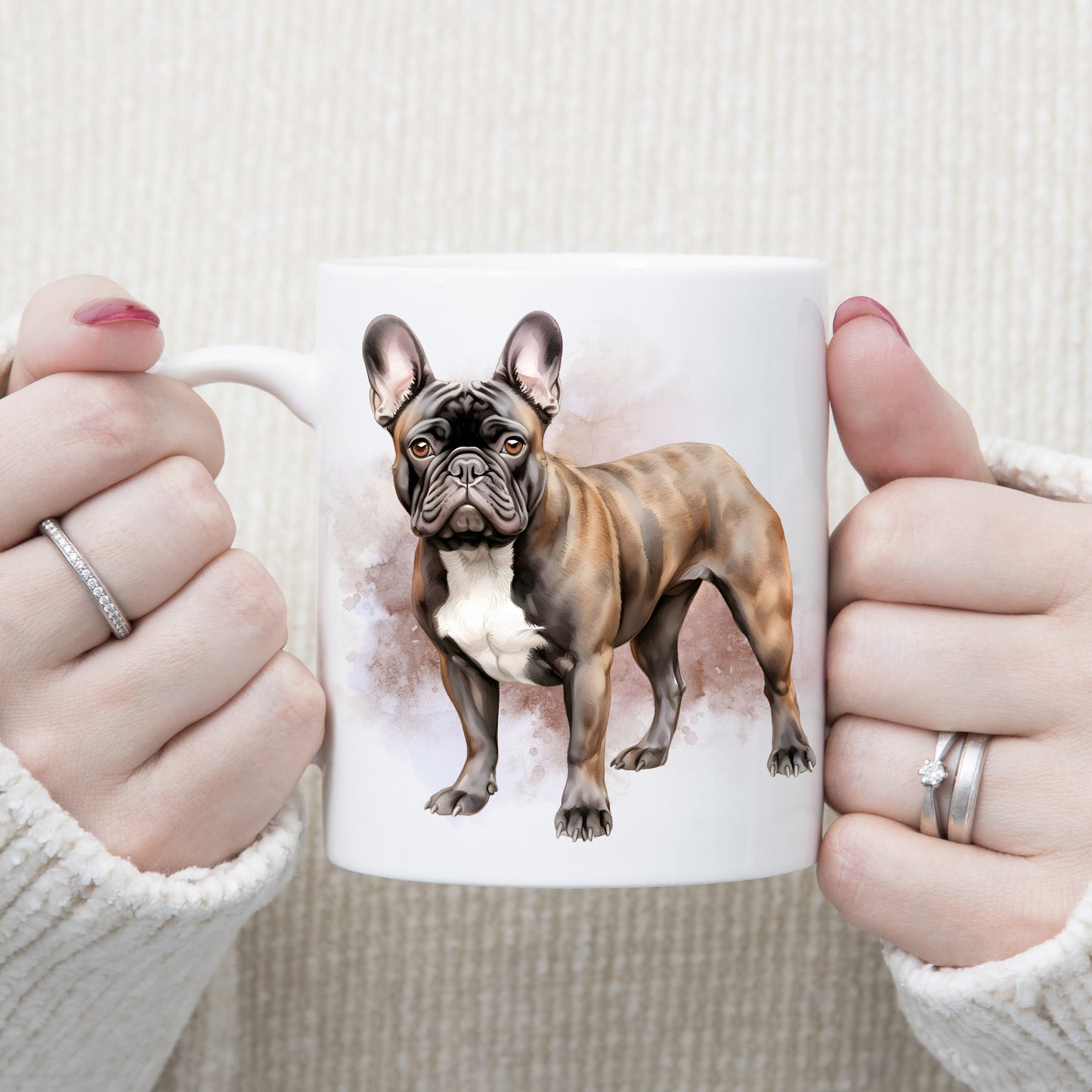 A watercolour image of a Brindle French Bulldog with a white chest adorns this white ceramic mug. A pink and brown misty background decorates the mug which is being held by a woman with both hands.