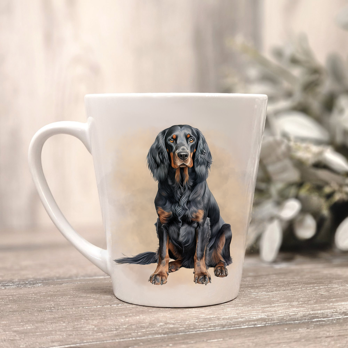 A seated Gordon Setter on a white ceramic latte mug in a smoky background and looking forward. The mug is placed on a wooden surface with sage green foliage to the right-hand side.