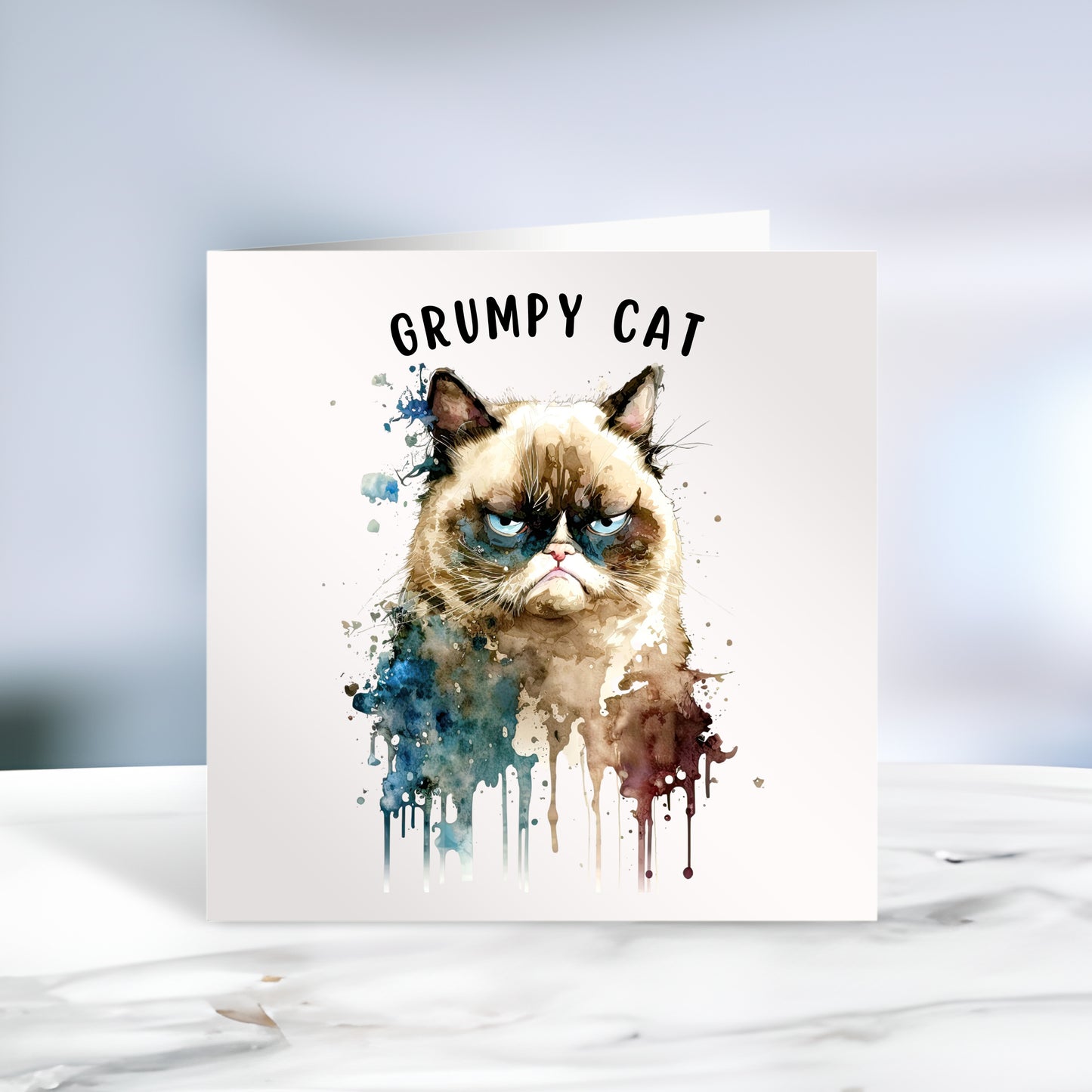 Grumpy cat on a white card with the words Grumpy Cat written at the top. Splashes and paint runs decorate. The card is placed on grey marble surface.