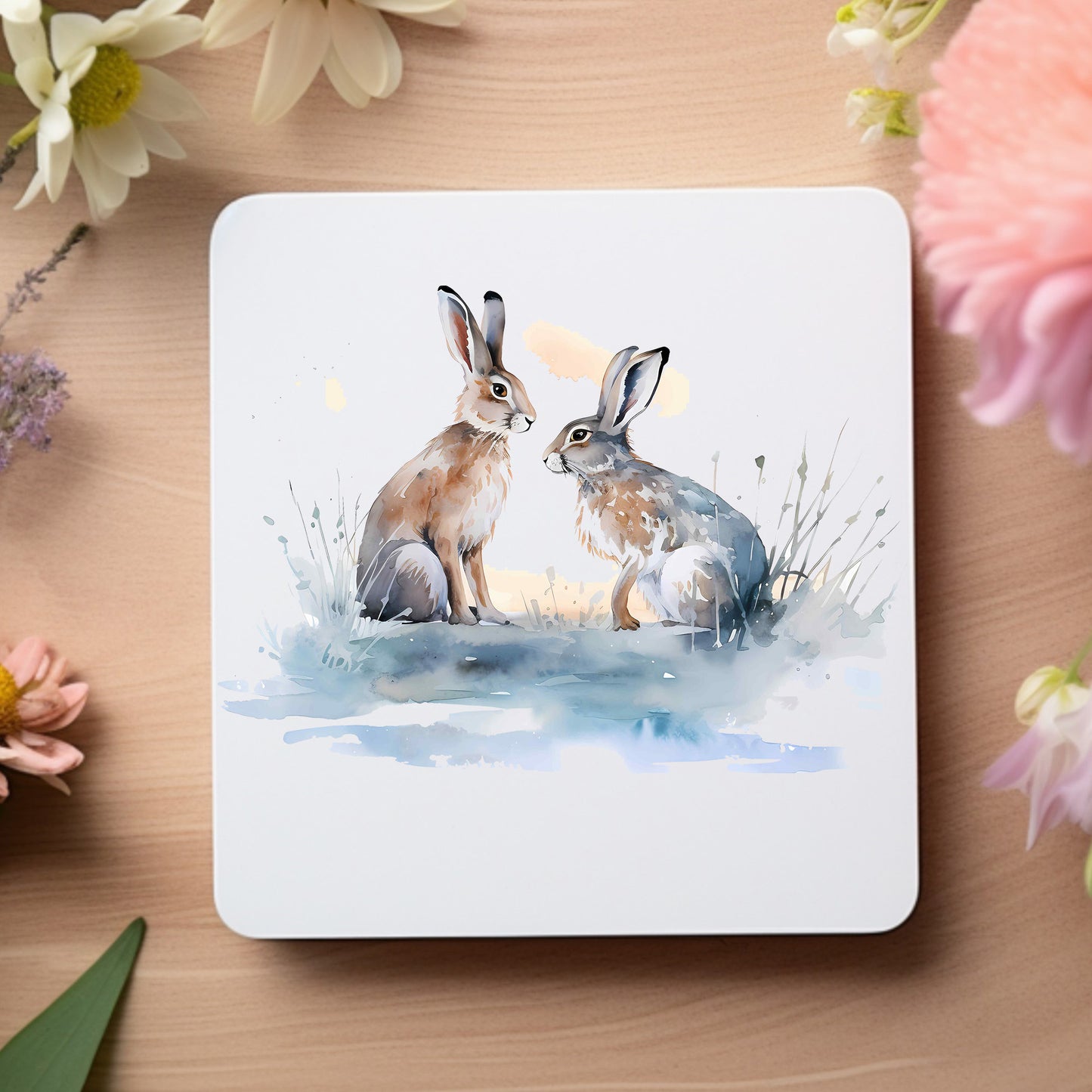 A pair of Winter Hares sit together in a snowy winter scene on a white coaster. The coaster is placed on a wooden surface flowers decorating the outer edge.