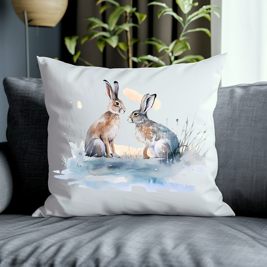 Two Hares face one another in a snow scene on the front of a white cushion which is placed on a grey sofa. A green plant decorates the background.
