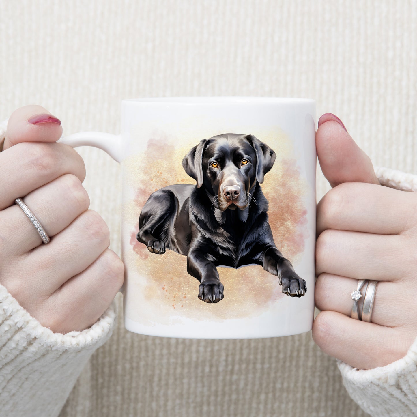A black Labrador dog is laid among a pink and gold misty background on a white ceramic mug. A woman is holding the mug