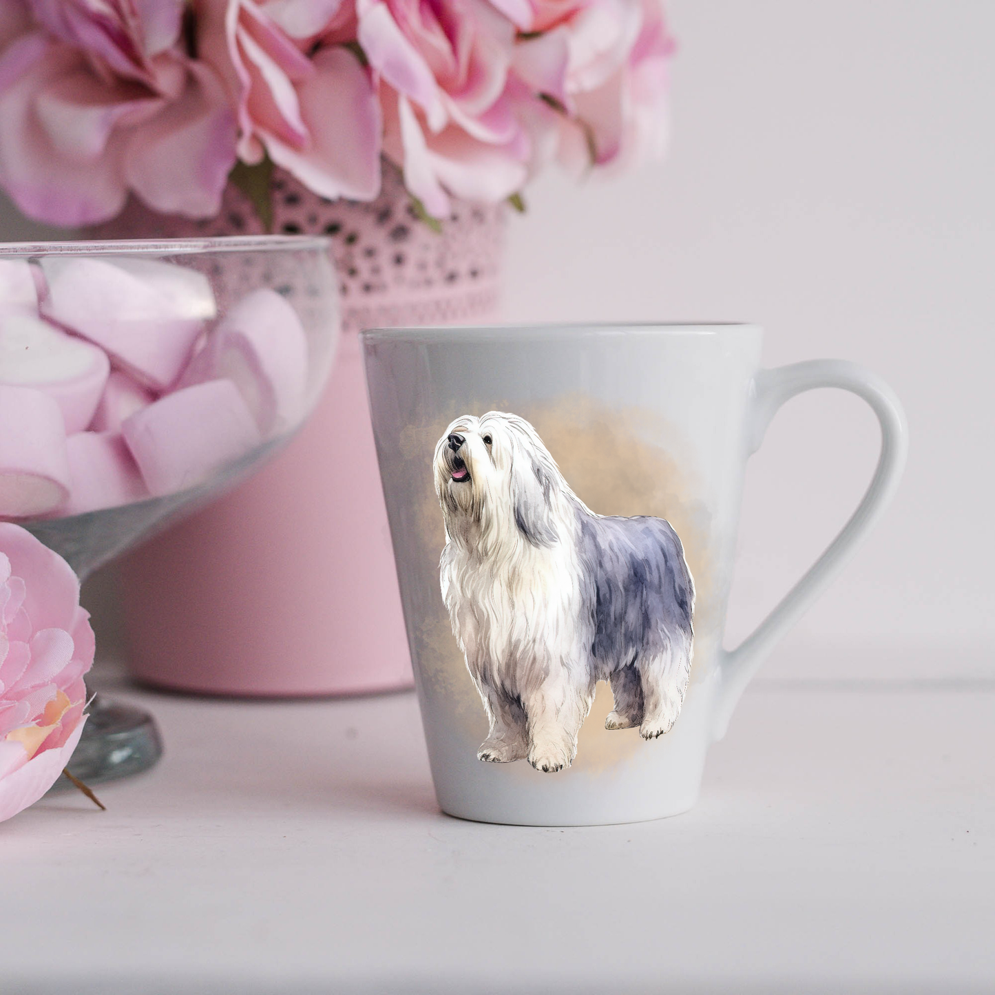 An Old English Sheepdog is seen standing on a white ceramic latte mug in a smoky background and looking forward. The mug is placed on a white surface with pink flowers in a pink plant pot and a dish of pink marshmallows.