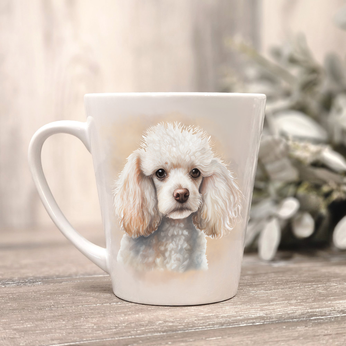 A white Miniature Poodle is shown on a white ceramic latte mug in a smoky background and looking forward. The mug is placed on a wooden surface with sage green foliage to the right-hand side.