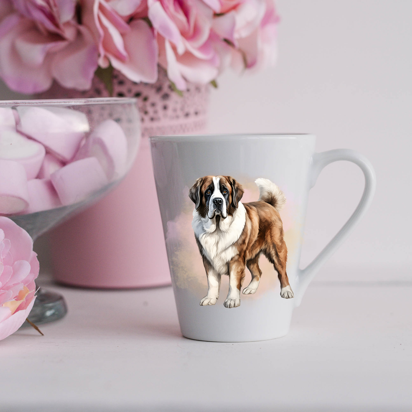 A Saint Bernard is shown standing on a white ceramic latte mug in a smoky background and looking forward. The mug is placed on a white surface with pink flowers in a pink plant pot and a dish of pink marshmallows.