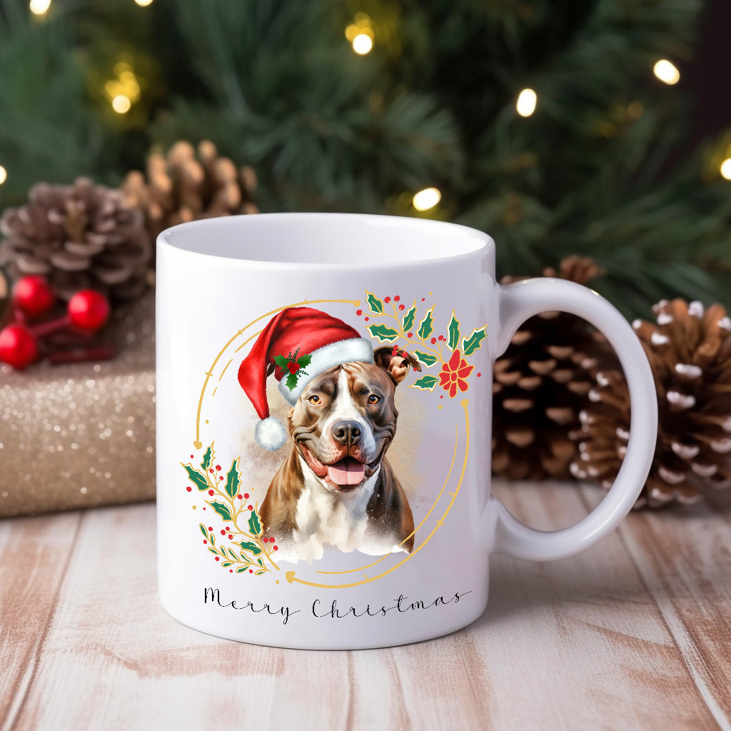 Staffordshire Bull Terrier wearing aChristmas hat on a Christmas themed mug. Placed on a wooden surface with pine cones and gifts behind.
