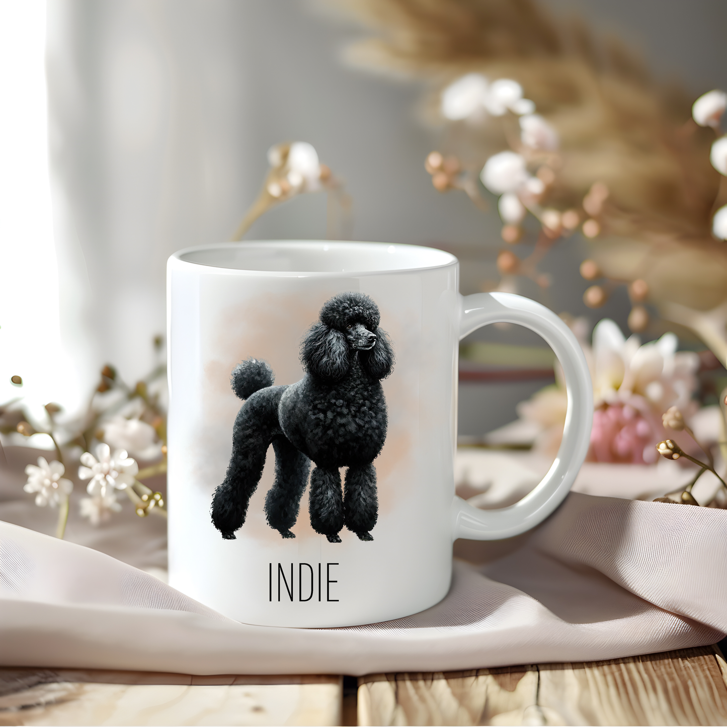 A lovely black Standard Poodle on a coffee mug personalised with a pet name. The mug is sat on a wooden surface with a creamy coloured cloth and a variety of flowers decorate the background.