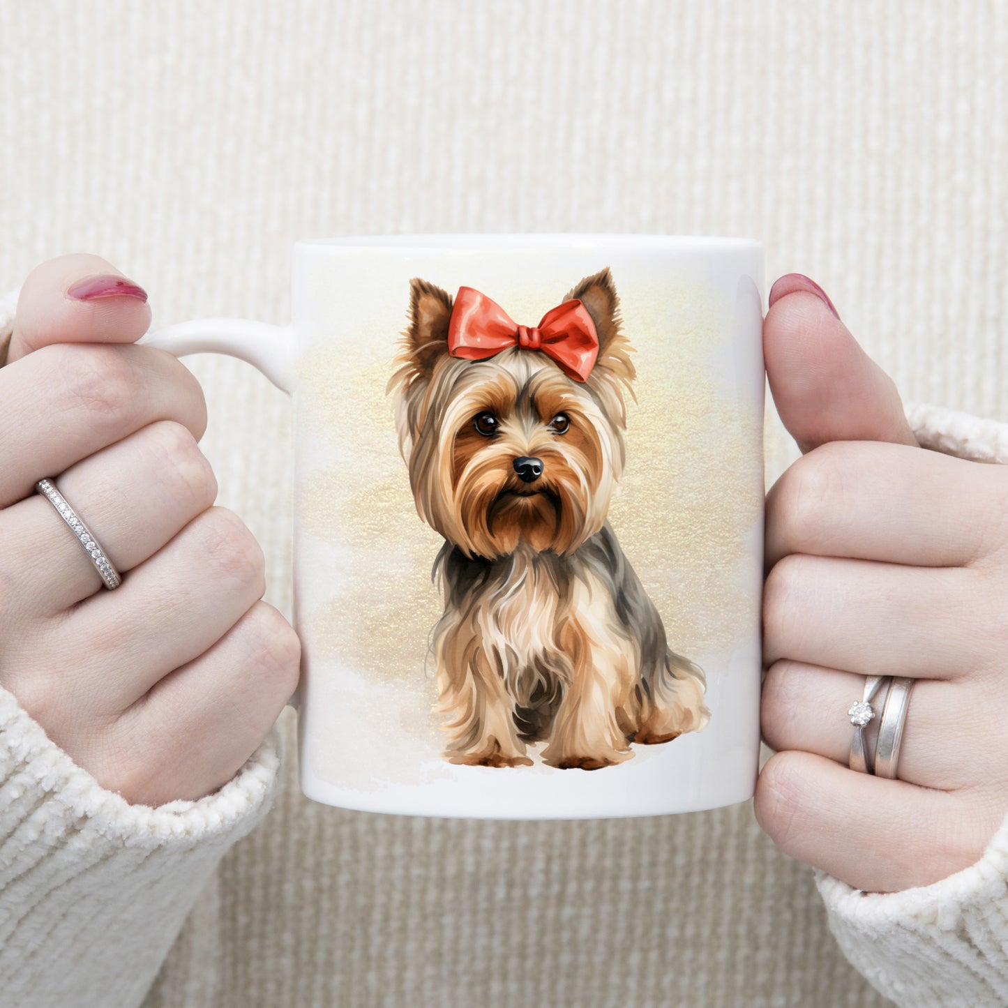 A white ceramic mug with a Yorkshire Terrier wearing a red ribbon bow. A gold and brown misty background decorates. A woman is holding the mug.