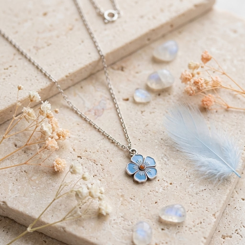 A silver-tone chain necklace with a small five-petal flower pendant featuring light blue enamel and a tiny gold centre, styled on a beige stone slab with dried flowers, moonstone beads, and a pale blue feather.