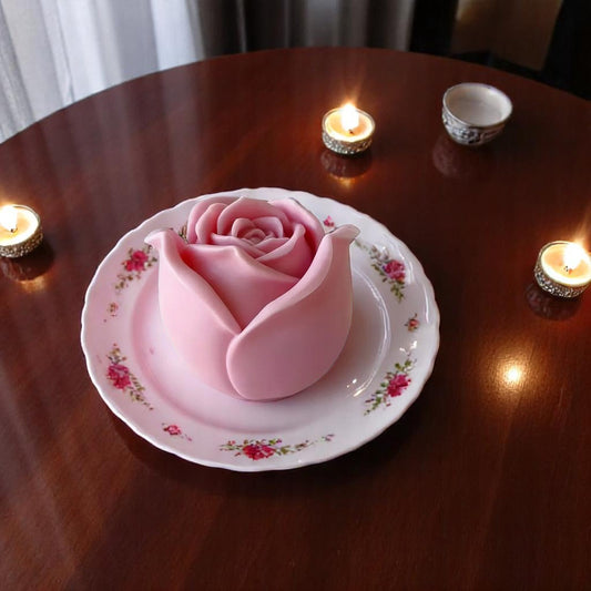 A pink wax melt shaped like a rose, placed on a plate with a floral pattern, surrounded by lit tea lights on a wooden surface.