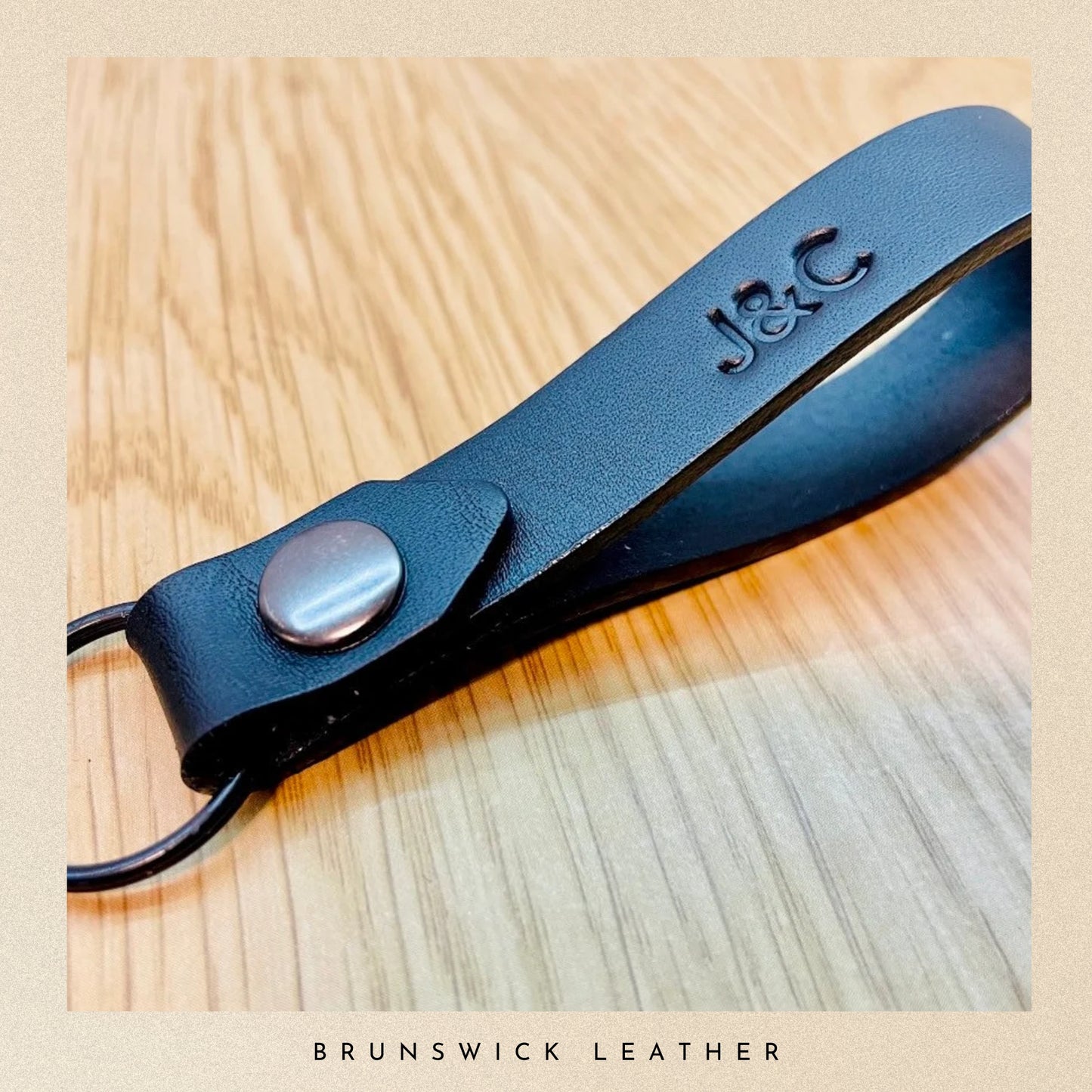 Black leather strap with a button on a wooden surface, featuring 'J&C' branding.
