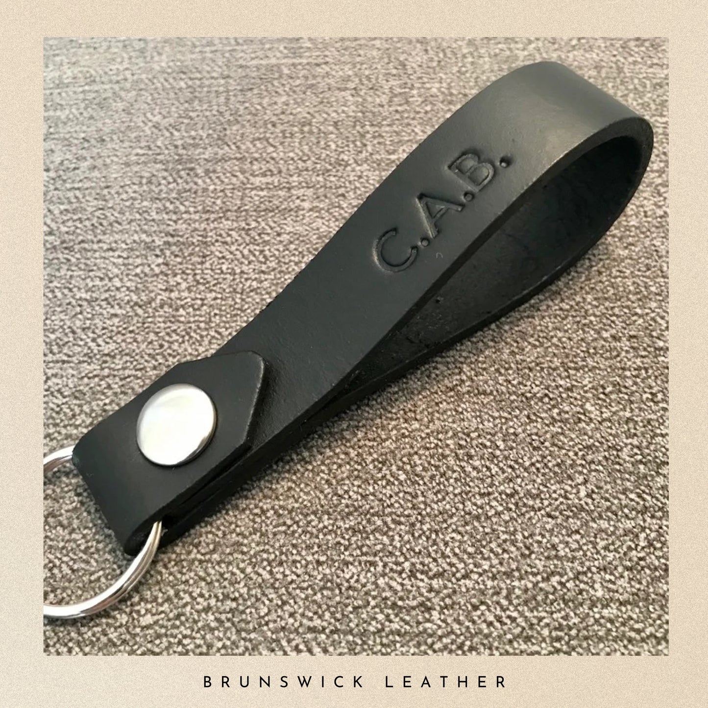 Black leather keychain with 'C.A.B.' engraving on a textured beige background, branded 'Brunswick Leather'.