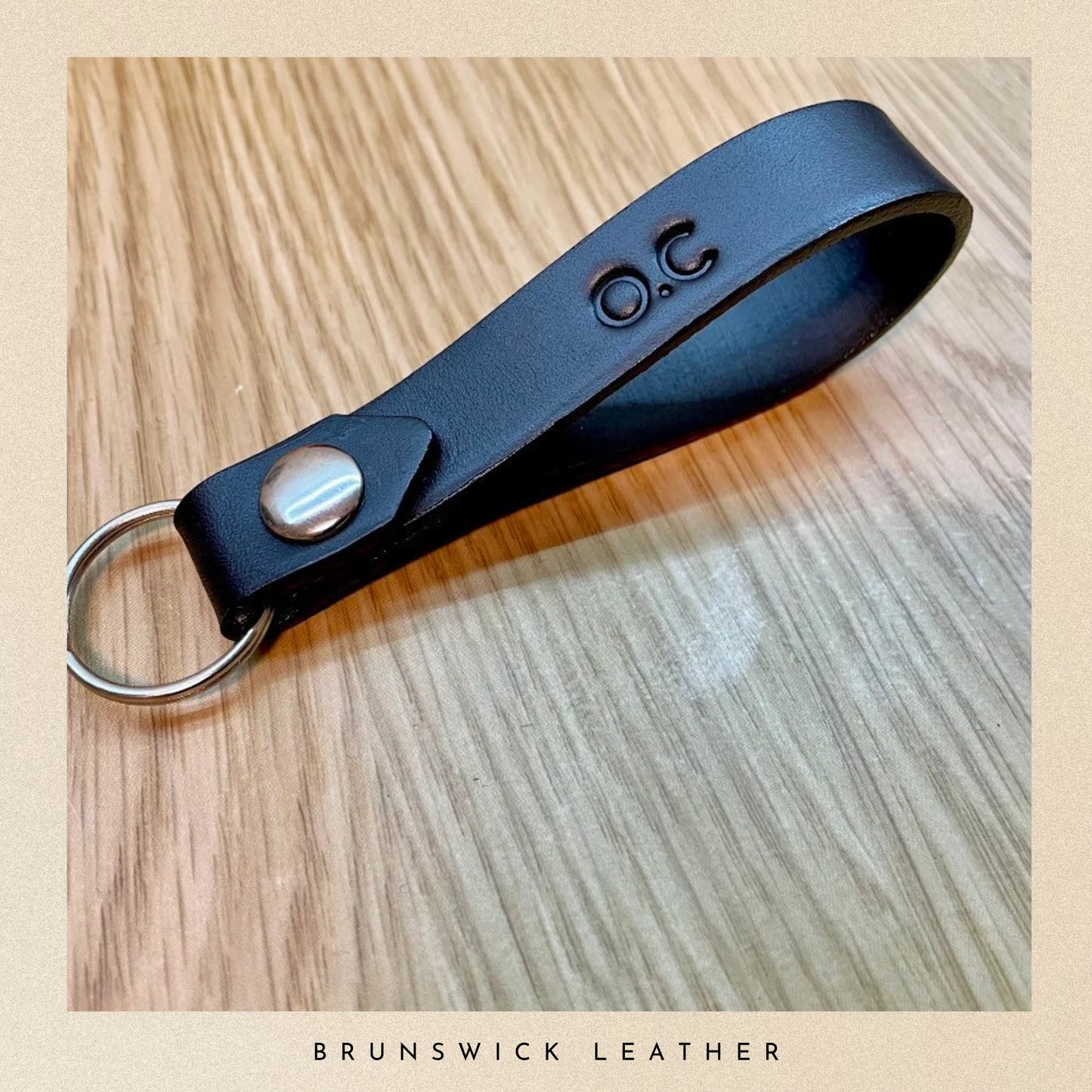 Black leather keychain with metal ring on a wooden surface, branded 'Brunswick Leather'.