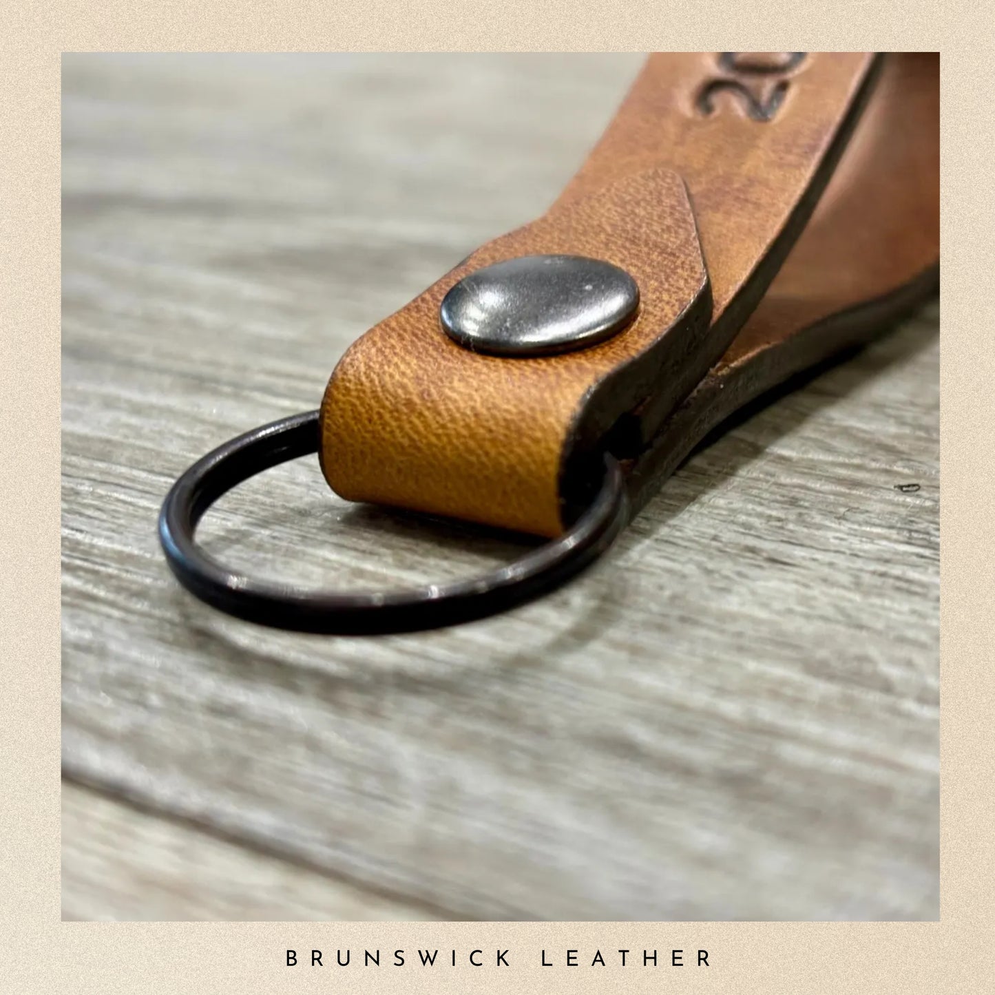 Brown leather keychain with metal ring on a wooden surface, branded 'Brunswick Leather'.
