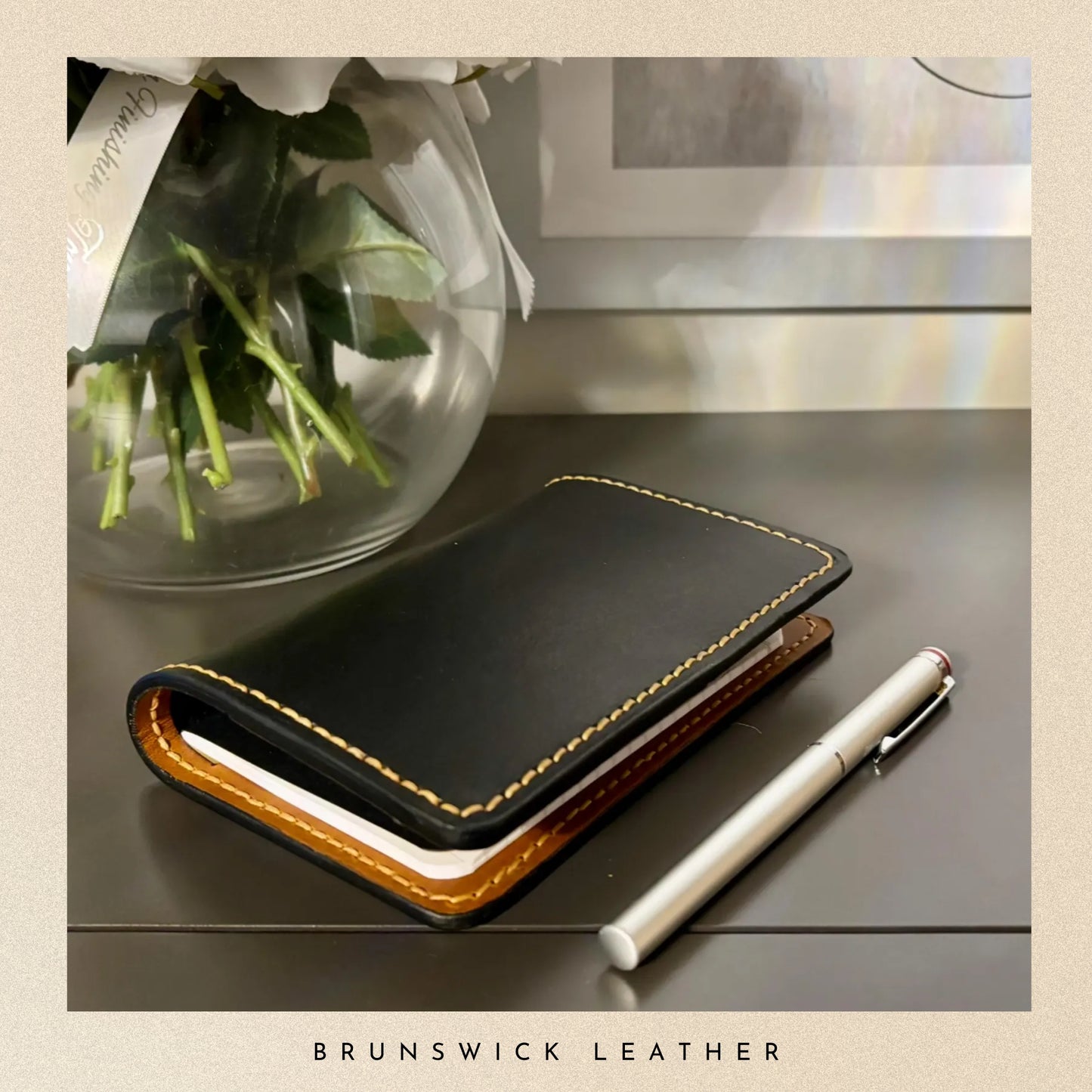 Black leather notebook with gold stitching on a wooden surface, next to a white pen and a vase with flowers.
