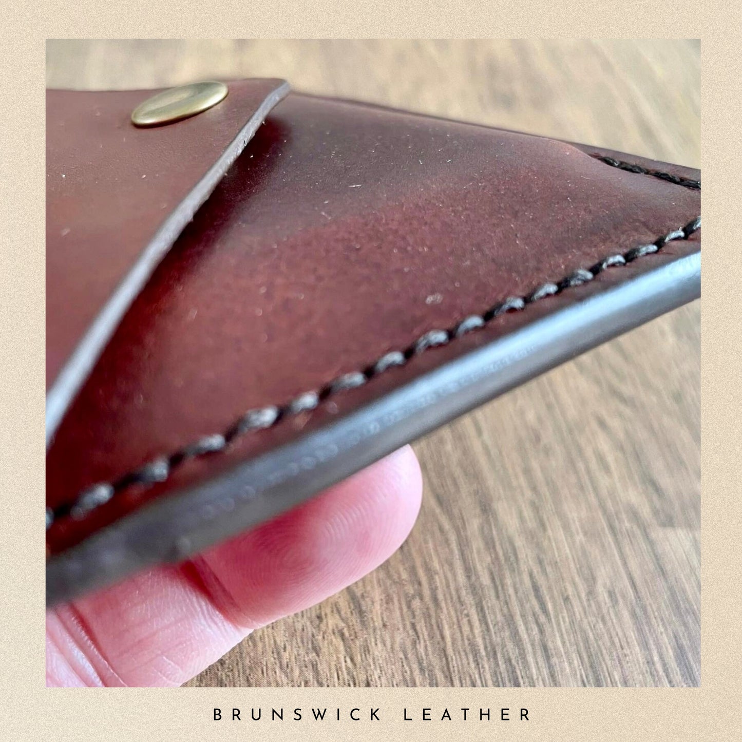 Close-up of a brown leather wallet held by a hand on a wooden surface, with 'Brunswick Leather' branding.