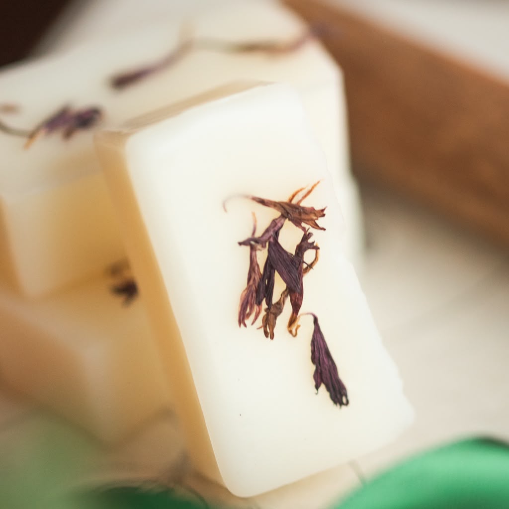 A close-up of a rectangular bar of off-white wax melts with dried dark purple flower petals embedded on its surface, with additional soap bars blurred in the background.