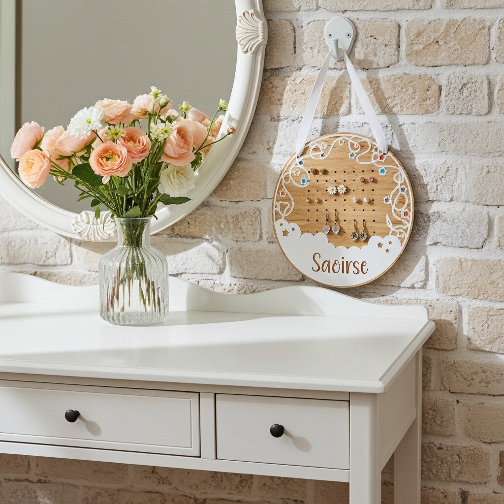 A round wooden jewellery display board hanging on a white wall hook above a white vanity table, with peach roses in a glass vase reflected in an oval mirror. The board has decorative white scrollwork, small clear gem accents, and the name 'Saoirse' printed at the bottom.
