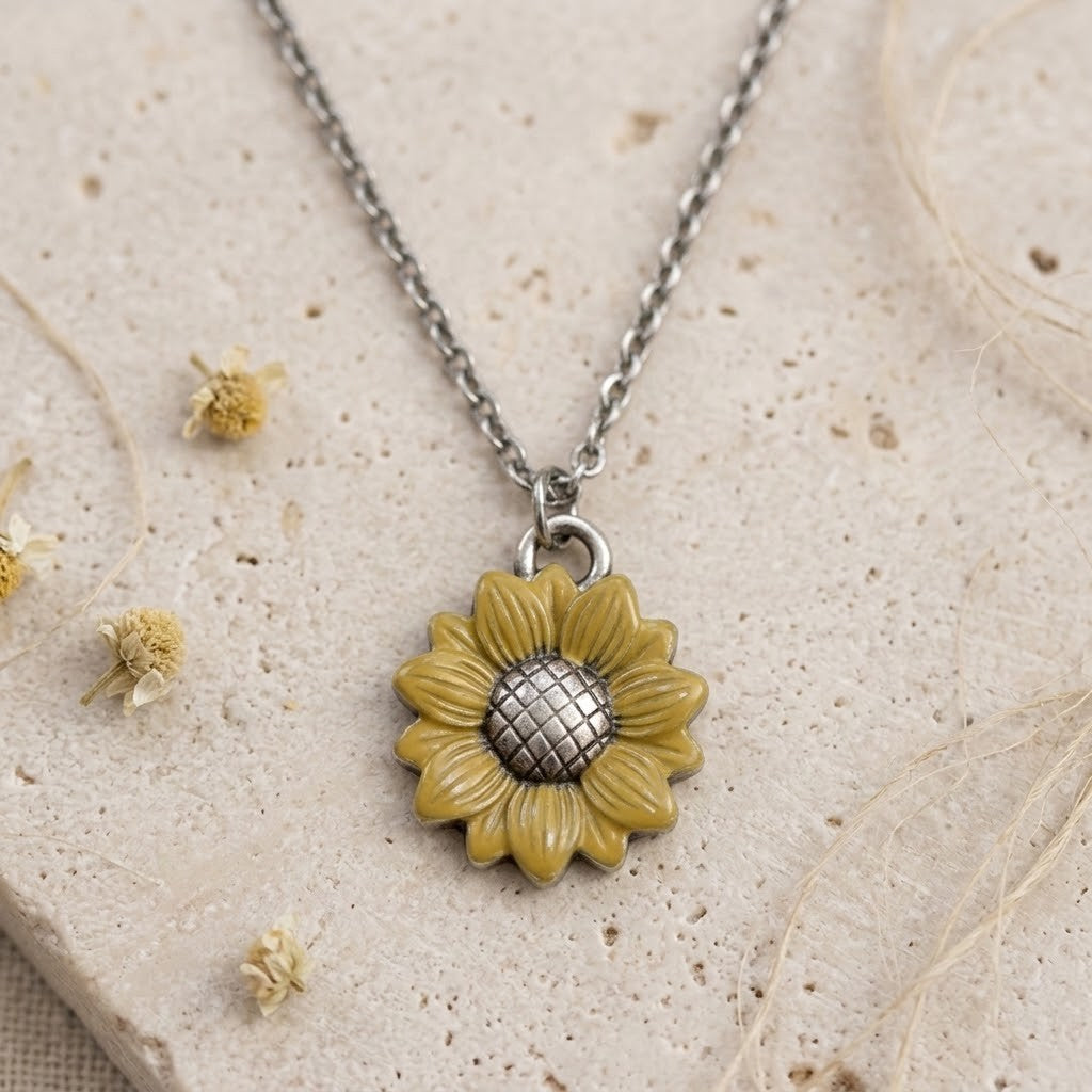 A sunflower pendant necklace with a textured yellow-green flower and a silver grid center, hanging from a silver chain on a beige stone surface with dried flowers.
