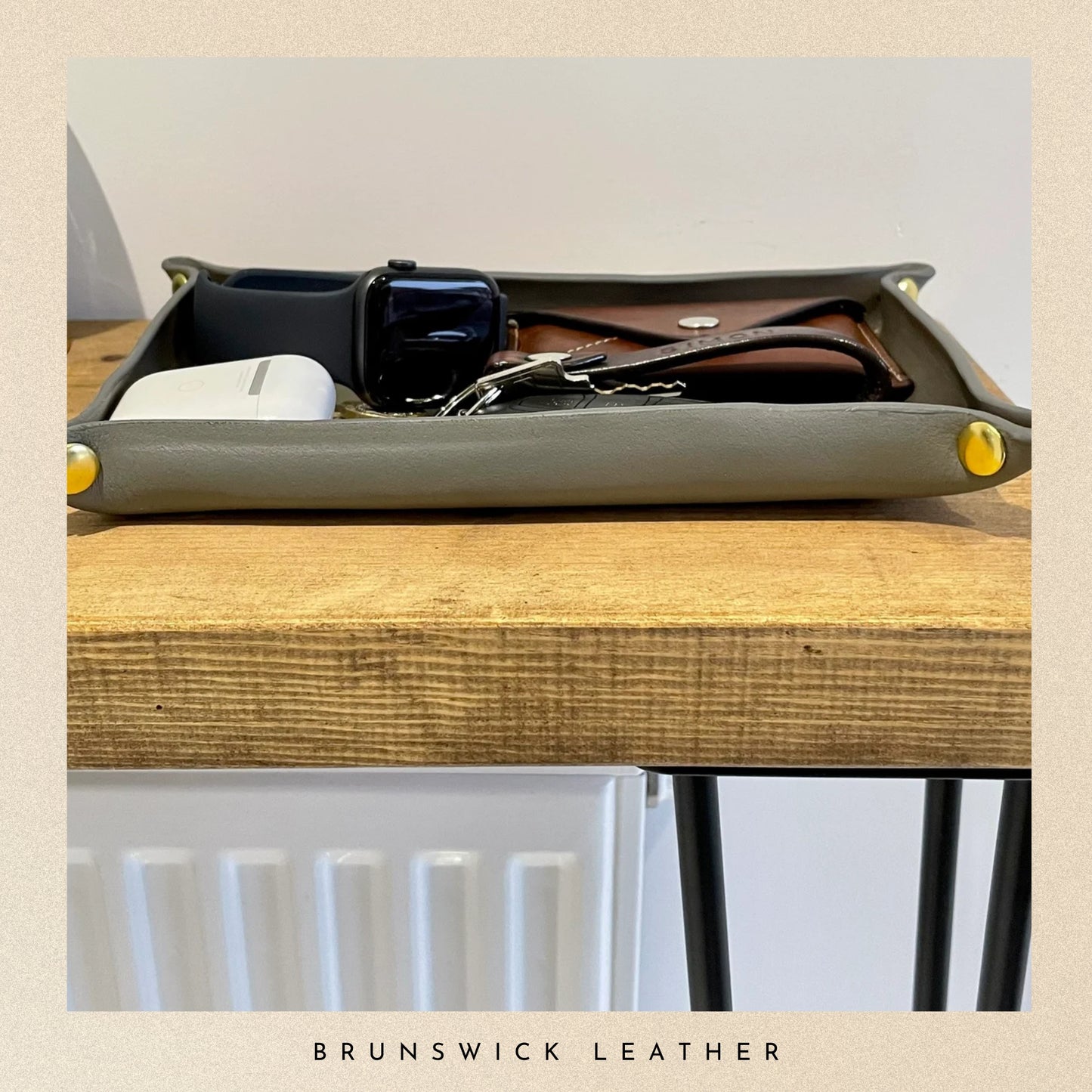 Gray leather tray with small items on a wooden surface, branded 'Brunswick Leather'.
