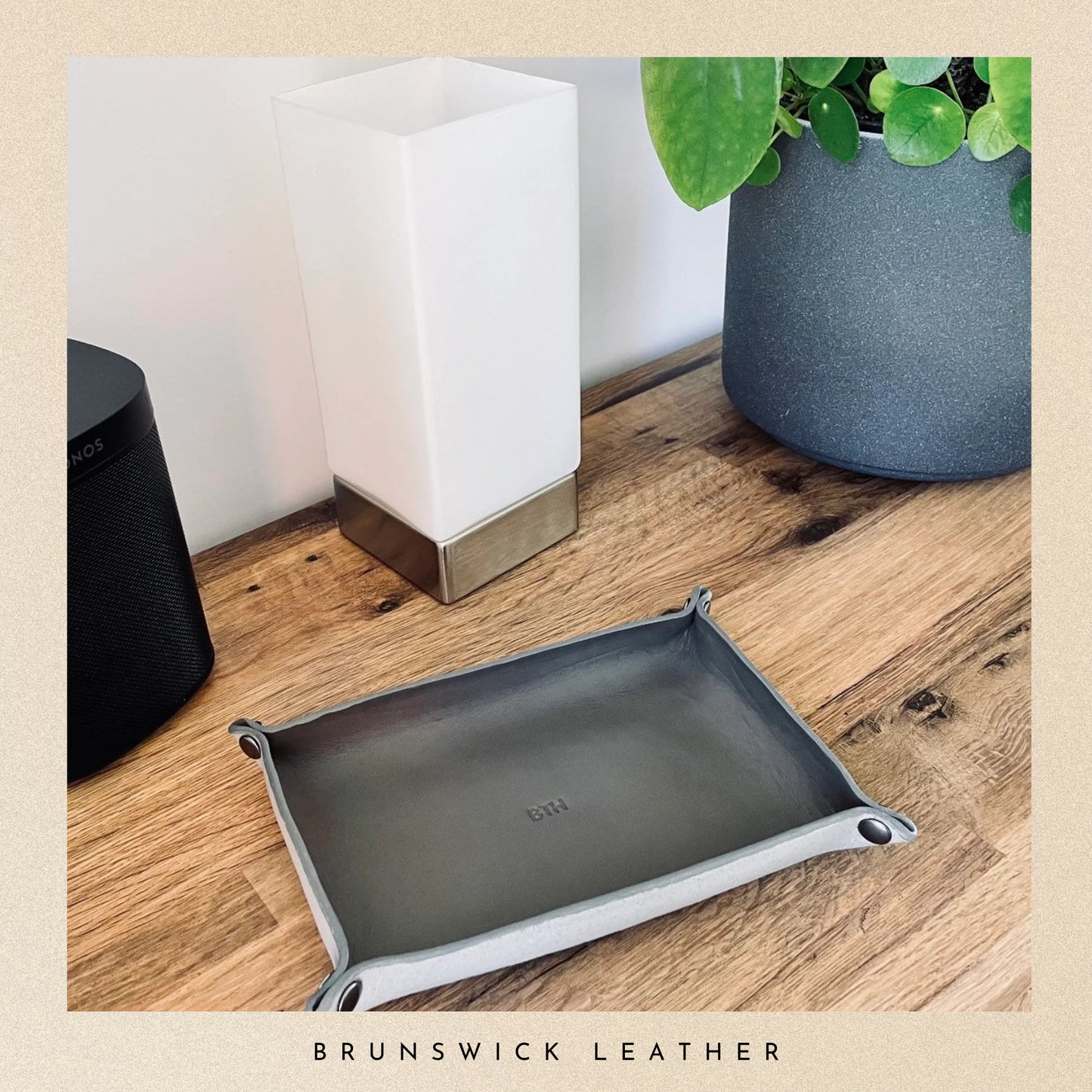 Grey leather tray on a wooden surface with a lamp and plant in the background, branded 'Brunswick Leather'.