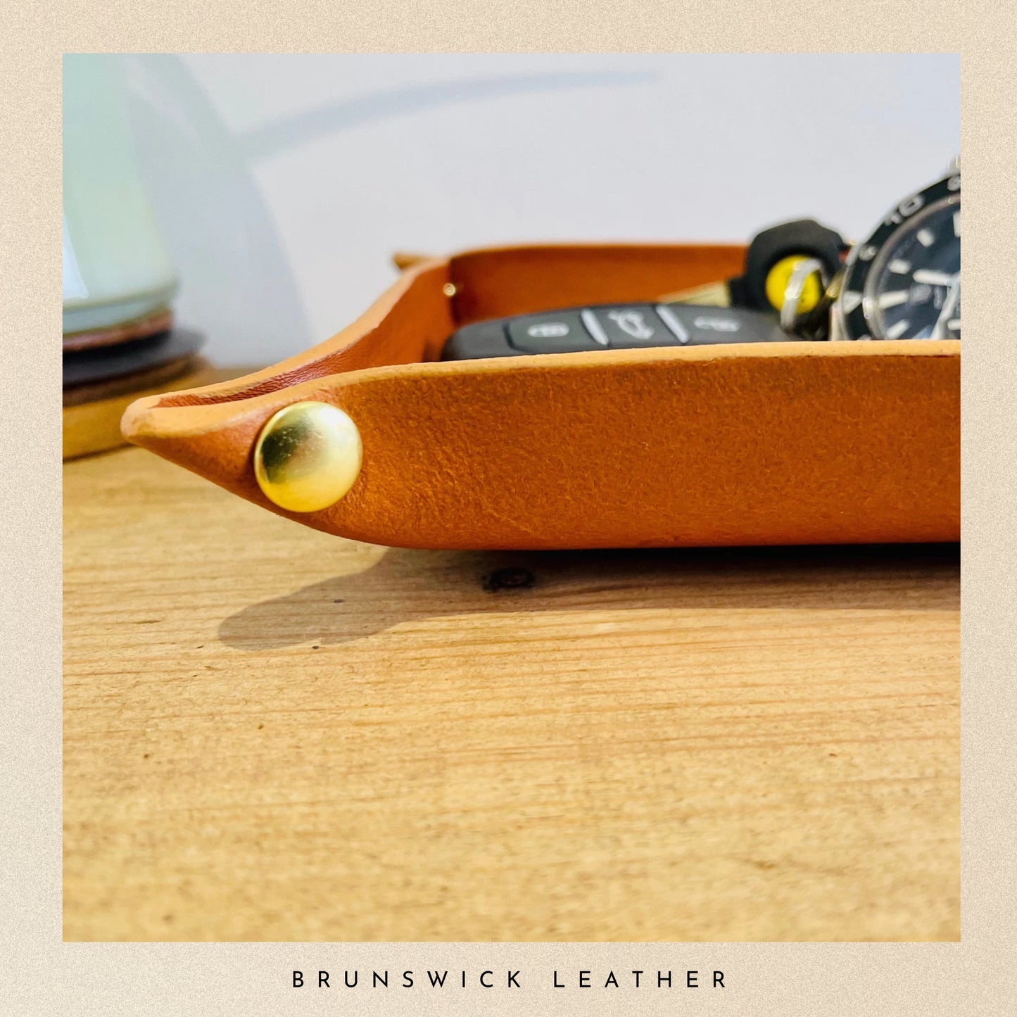 Brown leather tray with gold button on a wooden surface, featuring a watch and remote control.