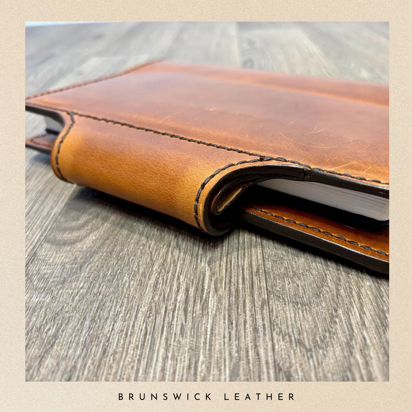 Close-up of a brown leather notebook with visible stitching on a wooden surface, branded 'Brunswick Leather'.