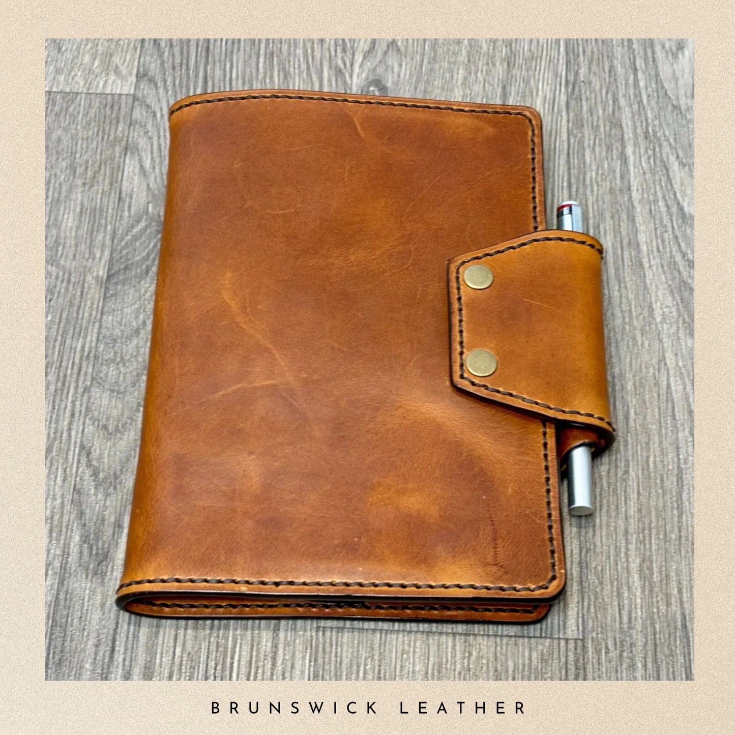 Brown leather notebook with pen holder on a wooden surface, branded 'Brunswick Leather'.